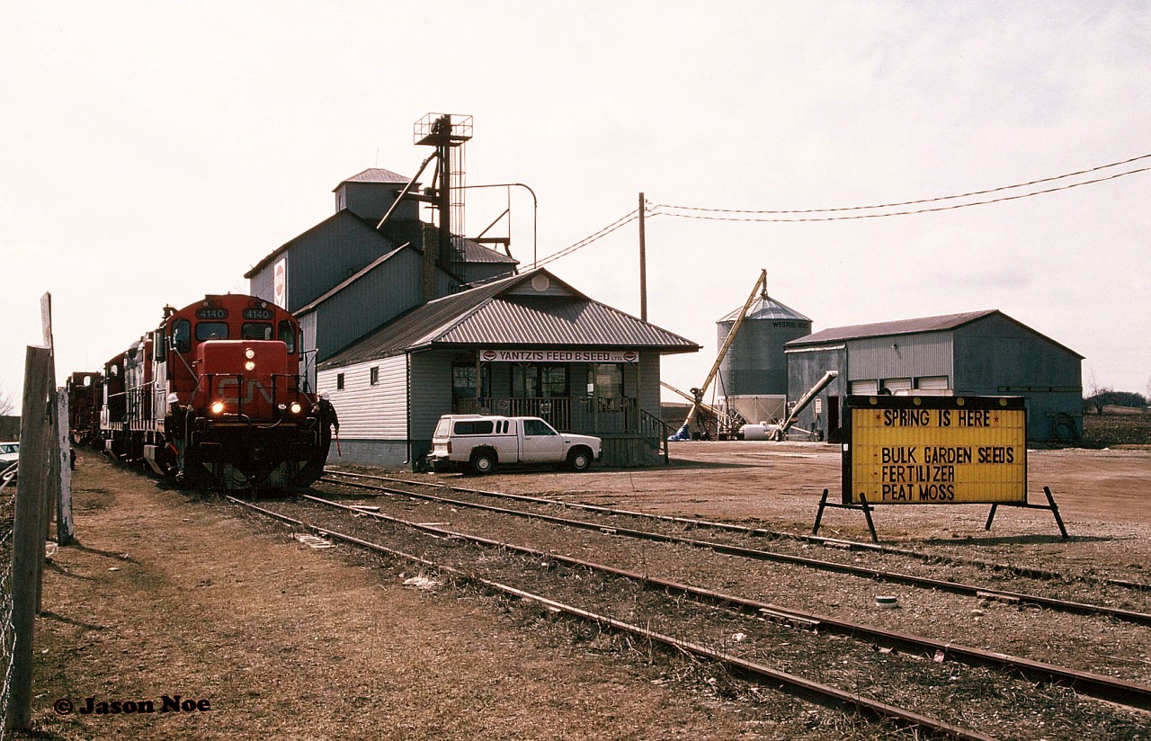Brunner is a small community in the Township of Perth East and was situated at Mile 11.5 of CN’s Newton Subdivision in Perth County, Ontario.
Spring is in fact here during April 1996 as CN 4140 and 4115 slowly pass the Yantzi Feed & Seed facility in the heart of Brunner as they haul the empty rail train towards Palmerston on the Newton Subdivision with the final destination of the town of Harriston. The train and its foremen are preparing to flag Highway #19, which is the main road through the town.
Today if you look on Google Maps street view more than two decades later, this area looks surprisingly similar, minus the railway of course.