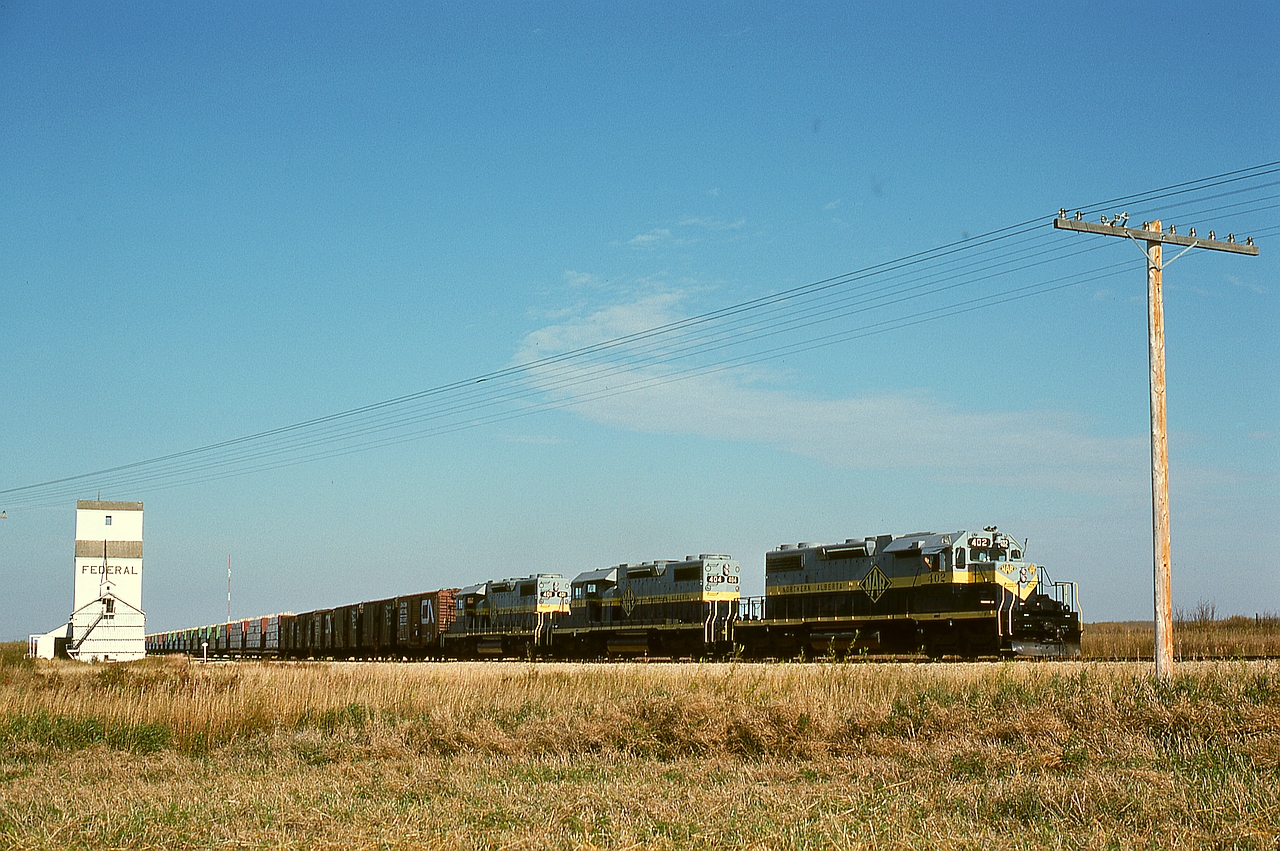 Under a glorious Peace River country clear sky on Friday 1976-09-24, Northern Alberta Railways’ GSL Turn is returning from the CN Great Slave Lake line connection at Roma Jct. to McLennan with its usual trio of SD38-2s, this day 402 plus 404 plus 401, passing the lone grain elevator at Judah at mileage 41.6 after the 8-mile climb from Peace River, where each unit was rated to handle 1200 tons.  In another 43 miles they will be at their home terminal.