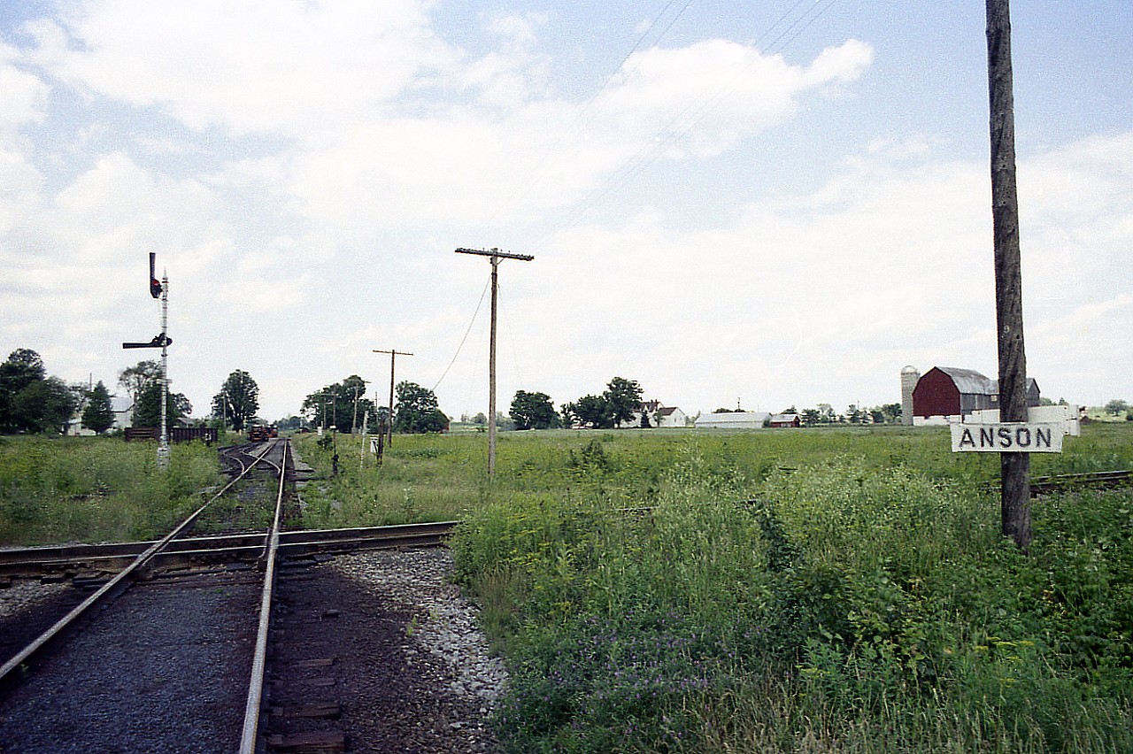 About halfway between Stirling and Hoards; maybe better posted as about 3 miles west of Stirling, lies this former diamond.  At this location the CN Campbellford Sub crosses the CN Marmora Sub, which is in this photo the line running Top to Bottom.  The location was but a crossing of the two routes, with a connector and siding on the northwest side; as one can see a gon on the connector and some MoW farther along. And a tool shed sort of building up by the road crossing, distant, on the right.
The location was referred to as Anson Jct by the railroaders from the old Central Ontario Railway that first put down the line thru here in 1883.
On the lower right where the sign is situated I believe is where a small frame building once stood, it was used by the crews during train operations when no one was on duty there. Too small to be a station so it was not much more than a railway operations point.  I'm wondering if the building in the distance is the same one that once stood on this spot.
Both lines were taken up in the mid 1980s. Not much to look at, but this location was once busy and significant to railroading in eastern Ontario.