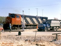 CN M420W 2516 handles a ballast hopper amid construction going on for the St. Clair Avenue East grade separation in Scarborough, as a worker stops traffic to direct a GMC dump truck working on the site. This is likely the temporary road diversion crossing installed next to the actual crossing (closed and to be ripped up for the grade separation work), which could explain the new crossing signals installed but not yet active (no crossing gates yet!)<br><br>This busy 4-track grade crossing next to GO's Scarborough Station on CN's Kingston Sub mainline was the site of a fatal collision in December 1975 between a TTC bus that got stuck on the crossing (due to issues with a faulty rear door interlock) and an eastbound Lakeshore line GO train running express. Part of the aftermath was a stronger push for a grade separation here, construction of which began two years later in 1977. Apparently the location had been earmarked for a grade separation for years, but kept being put off. Judging by the proximity of Midland Avenue (also grade separated at the same time, see 1977 view) and the junction between the CN Kingston and Uxbridge Subs in the area, a grade separation here was probably considered an extensive project.<br><br>See a <a href=http://jpeg2000.eloquent-systems.com/toronto.html?image=ser12/s0012_fl1970_it0095.jp2><b>before aerial view in 1970 </b></a> (grade crossing at bottom left) vs a <a href=http://jpeg2000.eloquent-systems.com/toronto.html?image=ser12/s0012_fl1977_it0040.jp2><b>during construction view in 1977</b></a>, vs an <a href=http://jpeg2000.eloquent-systems.com/toronto.html?image=ser12/s0012_fl1981_it0022.jp2><b>after aerial view in 1981</b></a><br><br><i>Mike Mastin photo, Dan Dell'Unto collection slide.</i>