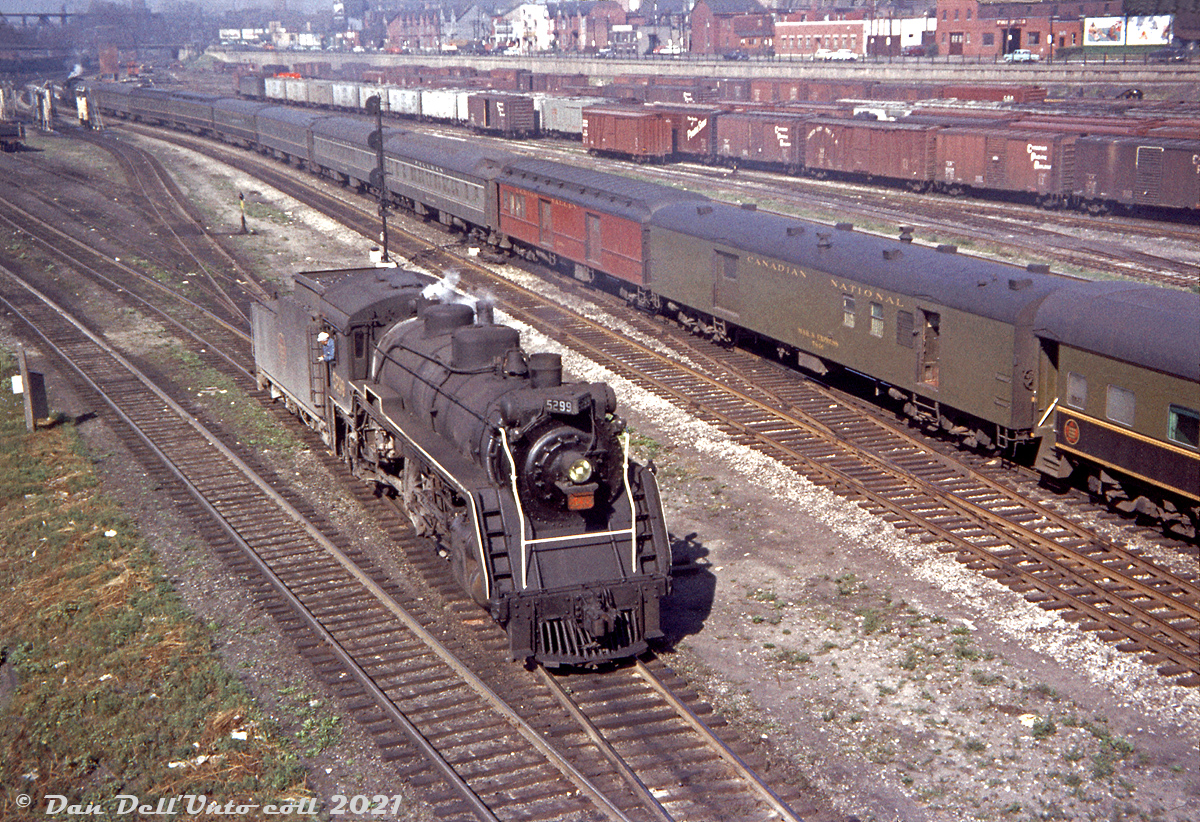 Canadian National 5299, a J7c-class Pacific (built by MLW in July 1920, scrapped March 1959) assigned to passenger service out of Spadina Roundhouse is seen reversing onto one of the yard leads near the engine servicing facilities downtown, looking west off Spadina Avenue sometime in the mid-late 1950's. CN's Spadina Coachyard is located to left out of view, its wash racks visible in the distance.

Two tracks over on one of the main tracks leading from Union Station, a CN steam engine pulls a passenger train with the usual CN-painted equipment, plus some NYC-painted Pullman sleepers, a Lehigh Valley RPO, and a CN RPO (lettered "Mail & Express").

Further back is CN's Bathurst St. Yard full of freight cars, predominantly a mix of 40' steel and wood boxcars: CN wafer logo cars, CPR stepped lettering cars (being interchanged with CN), a Northern Pacific outside-braced wooden boxcar, and a D&LW "Route of the Phoebe Snow" boxcar. A bunch of silver-grey CN 40' ice reefers are also present. One can just pick out the red farm equipment on flatcars coming from Massey Ferguson in Parkdale sitting near the west end. And in the upper right, a glimpse of the top of a CN diesel switcher hiding in the yard.

Buildings and houses along the elevated Front Street run along the background, meeting up with Bathurst St. and its bridge over the rail corridor on the upper left.

Original photographer unknown, Dan Dell'Unto collection slide (an unlabeled, undated and slightly faded Anscochrome that needed some TLC).