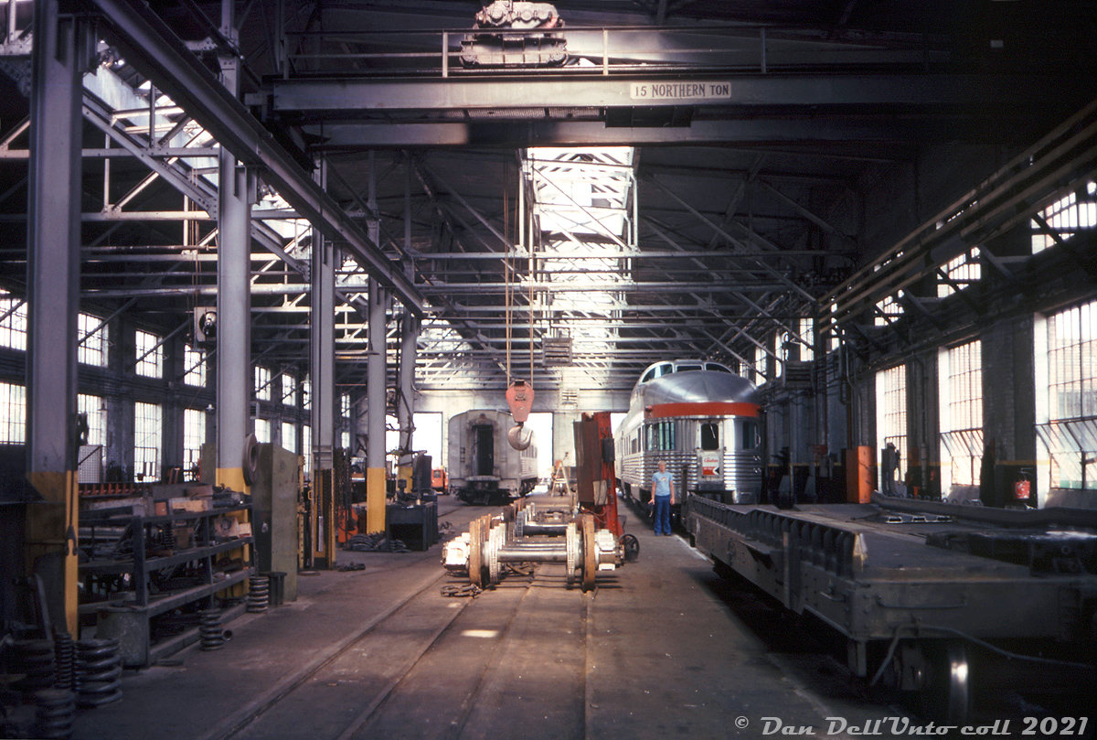 When one thinks of John Street they think of CP's famed roundhouse, that in later years maintained CP's fleet of passenger power, RDC's and later vintage Alco/MLW switchers until it closed in the mid-80's. But there were also other facilities on site at John Street: here's a rare look inside CP's 3-track John Street car shops building, located near the roundhouse just south of the CN tower, and used to maintain CP's fleet of passenger cars operating on trains out of downtown Toronto since it was constructed in the late 1920's for steam-era operations.

Three stainless steel Budd-built passenger cars used on The Canadian are in for work, including a "Park" series observation-dome-lounge car (likely Sibley Park). By this time in the mid-70's, service cuts and rationalization of CP's passenger network left few passenger trains to maintain out of Toronto. Trains #11/#12, the Toronto-Sudbury sections of "The Canadian" still called John Street home, along with the Peterborough and Buffalo Budd RDC runs. CP also operated piggyback ramps out of its John Street yards downtown starting in the 50's, and the carshops were also utilized to service CP's fleet of 50xxxx-series piggyback flatcars, like the one on the right and another barely visible on the left.

Down the middle between two tracks, passenger car wheelsets equipped with Budd's disc brakes and patented "Rolokron" anti-slip skid system are lined up in the rail indents between tracks below the 15-ton Northern overhead crane, ready for any change-outs. Truck springs of various sizes line the floor nearby, and a pile of brakeshoes can be seen further down.

From what I gather, what little passenger car servicing that remained here after VIA took over in 1978 was moved to VIA's ex-CN Spadina Coachyard at some point in the early-mid 80's (stainless steel ex-CP cars started appearing there before VIA's move to the TMC), and the car shops continued to be in use servicing freight equipment like piggyback and container flatcars (most piggyback operations had been moved to Obico, but nearby Parkdale Yard had been converted into a container handling facility) and storing work cars until John Street was shut down in July of 1986. The roundhouse was donated to the city by CP, but the car shops building was eventually demolished in the following years.

Bernie Smith photo, Dan Dell'Unto collection slide (from what I gather, that's Bernie's son in the photo, who made a few appearances in his photos).