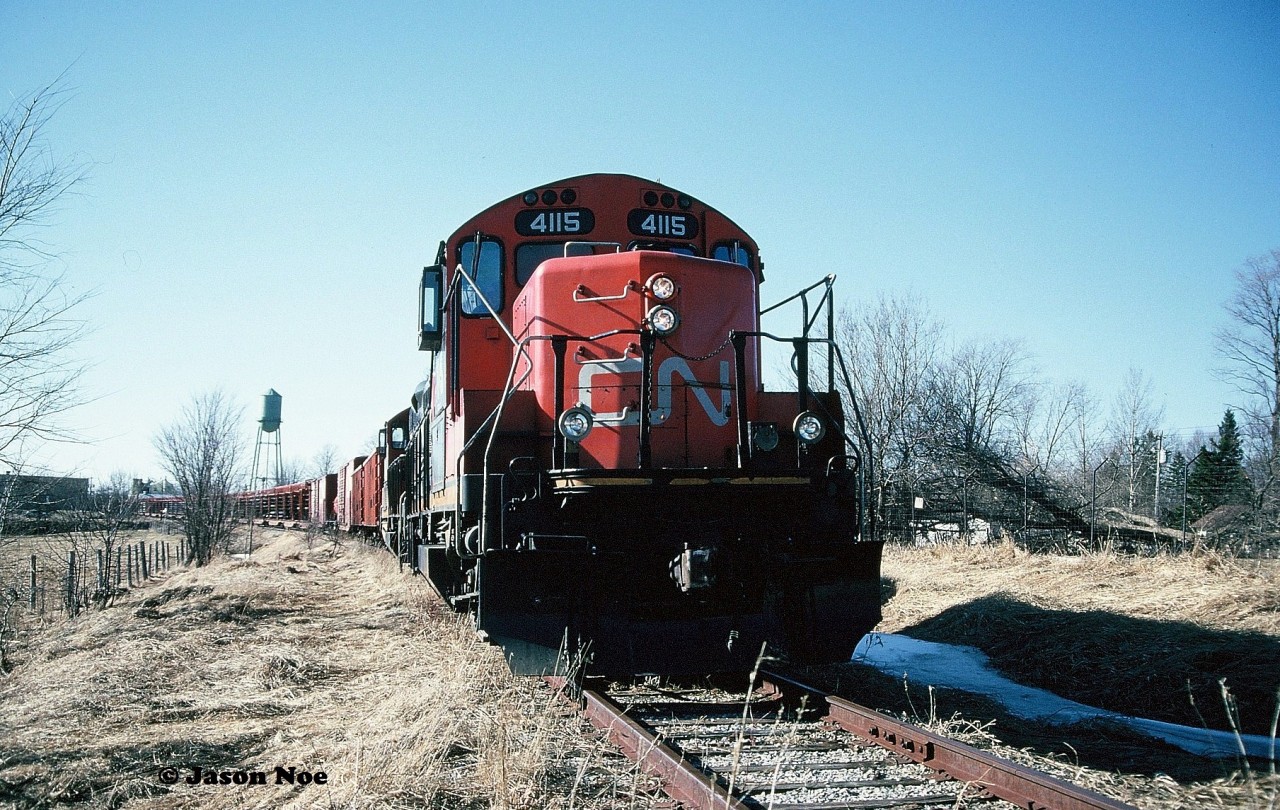 Pictured is a little better view showing the town of Harriston in the background with the rail salvage train. 

During the afternoon, GP9RM’s 4115 and 4140 with their rail salvage train have finally arrived in the town of Harriston and are seen awaiting instructions from the CN foremen accompanying the crew. The tail-end of the train was at the Raglan Street crossing as the town's old water tower can be seen to the left above the rail cars.