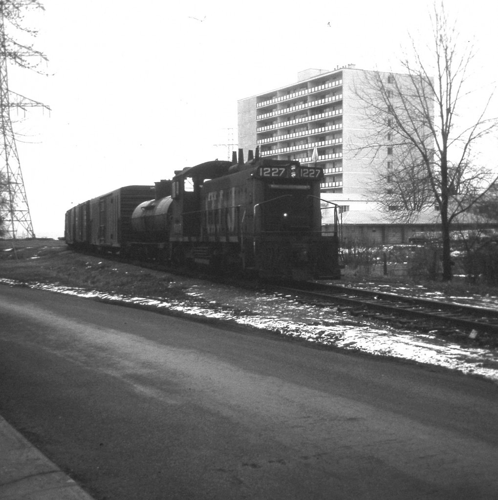 Dismal grey December day, but historical image. This view is of CN 1227 running eastward to CN Oakville sub where the old Beach line joins up at Burlington West. The end of the train is about to clear Lakeshore Rd and behind me is the crossing on Brock St.  The train is returning from switching out cars at businesses at the end of the line down by Langs Food Ltd. Before the Stoney Creek traffic circle was removed in the early 70s and the QEW thru there rebuilt, the CN line extended another 0.8 mile past Langs and joined the CN Grimsby sub in Stoney Creek, near Lake Av. My recollection of the former Beach Sub is a bit fuzzy. I believe service ended in 1977, track abandoned in 1981 and pulled up in 1983. The location of this photo can be identified by the white apartment which is still standing at the corner of Lakeshore Rd and Maple Av. In the left background was where the 'famous' Brant Inn stood until about 5 years before this photo was taken. The area is all built up now.