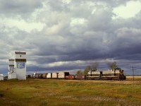 This return half of a Barrhead Turn is timetable eastward (compass southeast) on Northern Alberta Railways by the grain elevators at the hamlet of Mearns under an ominous sky on Saturday 1980-09-20, with GMD1s 312 and 303 for power and just 27 miles from home at Dunvegan Yards in Edmonton.  In the distance is the bell tower and spire of St. Charles Borromeo Catholic Church which, unlike those grain elevators, survives to this day.
