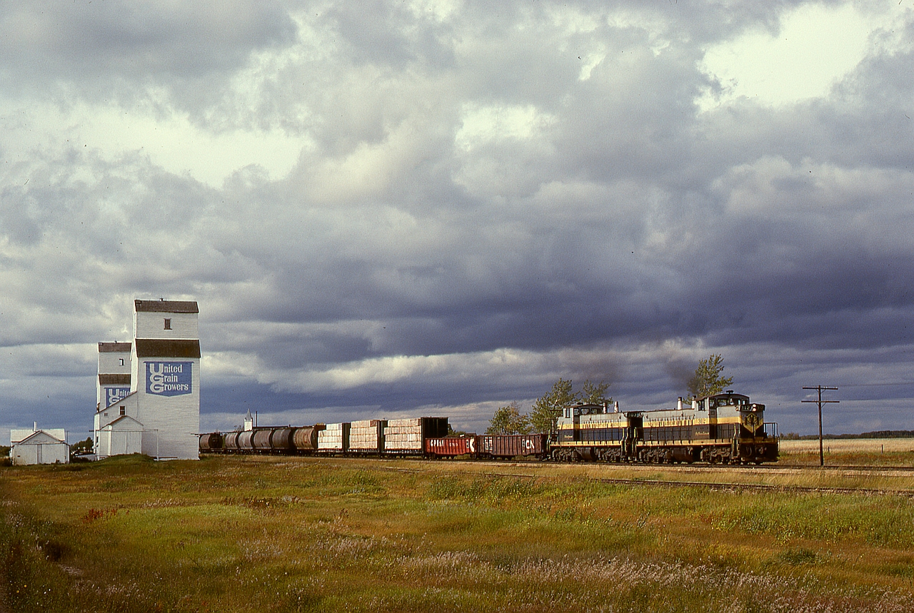 This return half of a Barrhead Turn is timetable eastward (compass southeast) on Northern Alberta Railways by the grain elevators at the hamlet of Mearns under an ominous sky on Saturday 1980-09-20, with GMD1s 312 and 303 for power and just 27 miles from home at Dunvegan Yards in Edmonton.  In the distance is the bell tower and spire of St. Charles Borromeo Catholic Church which, unlike those grain elevators, survives to this day.