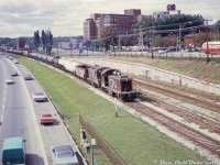 A typical Sunnyside scene: CN SW1200RS units 1219 and 1213 lead an eastbound freight (maybe a transfer from Mimico to Don or MacMillan Yard) past the old disused Sunnyside station platforms on a sunny afternoon. Traffic appears light with a number of cars pulled over on the shoulder for some reason, and two young spectators with their parents take in the lineside action. A TTC PCC (one of the 4725-4774 ex-Cleveland A11-class) heads east on the Queensway just outside Roncesvalles Carhouse (out of view to the right), with St. Joseph's Hospital looming in the background.
<br><br>
<i>Original photographer unknown, Dan Dell'Unto collection slide.</i>