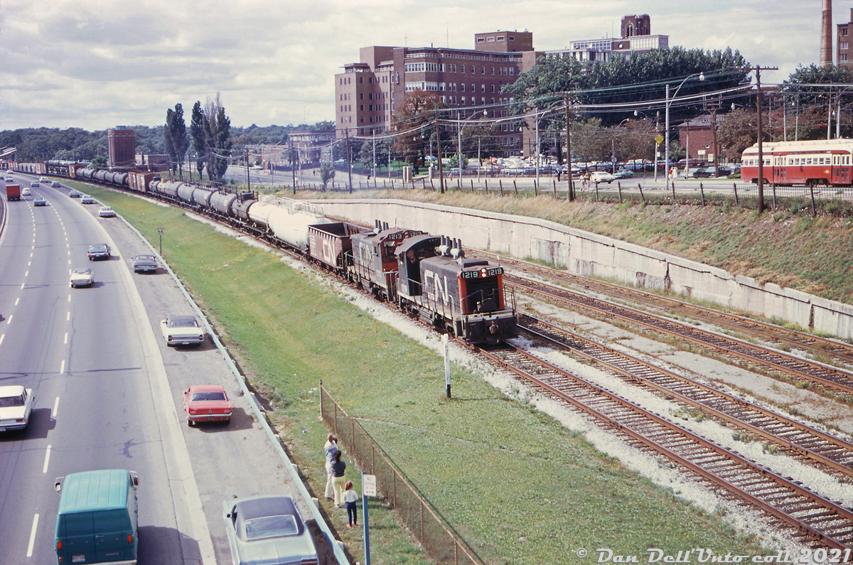 A typical Sunnyside scene: CN SW1200RS units 1219 and 1213 lead an eastbound freight (maybe a transfer from Mimico to Don or MacMillan Yard) past the old disused Sunnyside station platforms on a sunny afternoon. Traffic appears light with a number of cars pulled over on the shoulder for some reason, and two young spectators with their parents take in the lineside action. A TTC PCC (one of the 4725-4774 ex-Cleveland A11-class) heads east on the Queensway just outside Roncesvalles Carhouse (out of view to the right), with St. Joseph's Hospital looming in the background.

Original photographer unknown, Dan Dell'Unto collection slide.