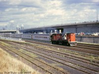 A westbound CN Tempo Train out of Union Station, lead by RS18 3150, speeds through the new Exhibition GO Station on the north passenger main track. Cars from annual fall Canadian National Exhibition patrons pack the parking lot between CN's Oakville Sub rail corridor and the Gardiner Expressway, including a farm tractor (probably not a regular GO commuter)! The top of the CNE's Ricoh Coliseum can be seen in the background. The double-wide pedestrian bridge to the north platform was built when the station opened, but by the late 90's was removed in favour of an underpass.<br><br>GO Transit's new Exhibition Station at this spot opened north of the CNE and south of Liberty Village sometime between September 1968 (after the 1968 CNE) and November 1968. The old CN steam-era Exhibition Station to the west at Dufferin Street was briefly used by GO in 1967-1968, but due to the poor setup of the old station handling large seasonal CNE passenger volumes, a new station was built to the east. At the time of this photo, the CNE still had a pair of rail siding of its own to the southeast of the new station for parking rail equipment.<br><br>CN Oakville Sub car control diagrams from 1970 show the two middle tracks referred to as passenger mainlines, and the two outer tracks as freight mainlines (presumably used by GO as well). At the bottom are storage tracks for a small CN Strachan yard, at the foot of the Liberty Village industrial area that was still full of sprawling spurs and sidings serving the likes of John Inglis, Canadian General Electric, and Toronto Carpet Manufacturing. This area was referred to in timetables as the "Jefferson Avenue District". Today of course, the area has been highly gentrified with new highrise condos mixed in with old industrial apartment/loft/condo conversions.<br><br><i>Original photographer unknown, Dan Dell'Unto collection slide.</i> 