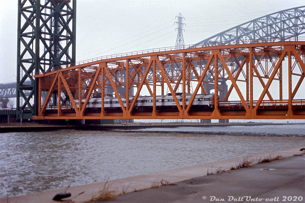 A rainy day down at the Burlington Beach lift bridge finds CN RDC-1's 6106 and 6111 crossing over the Burlington Bay Canal on CN's Beach Subdivision at Hargrove, likely doing a photo run-by for the Upper Canada Railway Society fantrip they're on. CN's Toronto to Niagara Falls Budd "Railiner" RDC runs typically operated through Hamilton via the Oakville-Grimsby Subdivisions, so a pair on the "Beach Branch" wasn't a common occurance. The QEW's Burlington Bay Skyway bridge is visible jut behind.  The CN Beach Sub from Burlington to Stoney Creek, originally a through-route for the Hamilton & North Western Railway, was soon severed due to mid-70's QEW freeway construction, and short spurs were operated at each end until abandonment in the early 80's. The two tracks that crossed the Burlington Canal lift bridge were replaced with extra traffic lanes.  Gord Taylor photo, Dan Dell'Unto collection slide.  Same UCRS fantrip at Stoney Creek: http://www.railpictures.ca/?attachment_id=43960 1974 Fantrip with GO power: http://www.railpictures.ca/?attachment_id=37233