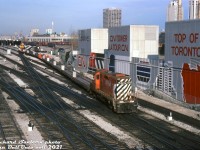 The 80's in Toronto was a transitional period regarding the presence of rail activity downtown, as both CN(VIA) and CP pulled out of their respective John Street and Spadina servicing facilities as local industrial traffic dried up, and passenger servicing was consolidated to more modern facilities out of the downtown core.<br><br>CP GP9u 1534, a relatively recent rebuild done by Angus Shops just a few years earlier (ex-8538), handles a transfer with a string of piggyback flatcars and old <a href=http://www.railpictures.ca/?attachment_id=46615><b>wooden van 436994</b></a> on the tail end, heading westbound through the Toronto Terminals Railway trackage by John Street interlocking.<br><br>The base of the CN Tower is on the right with an updated CN railway mural (no more Tempo and Turbo Trains the old version had). The CP John Street yard tracks in the background that once teamed with maroon and stainless steel passenger cars now only see old boxcars, OCS (On Company Service) work equipment, and piggyback and container flatcars stored. John Street would soon be closed in 1986, and the roundhouse proper donated to the City of Toronto.<br><br>The harbourfront skyline is relatively uncluttered here: the usual buildings are present including the Victory Soya Mills elevators, the Postal Building (real estate developers Bramalea Ltd & Trizec would buy it in the early 90's, but redevelopment fell through due to the recession. It eventually became home of the Air Canada Centre), the Toronto Star office tower at 1 Yonge St., and the Hilton Harbourcastle hotel.<br><br>The two smokestacks on the right were for the nearby Central Heating Plant the provided steam heat to downtown buildings including John Street Roundhouse, Union Station, and the Royal York Hotel. It would be demolished in 1990, around the time when the land around John St. was cleared for future redevelopment. As part of the project, the former yard area was excavated and part of the old York Street viaduct under the yard was demolished, shortening the viaduct to stretching just under Union Station's train shed.<br><br><i>Richard Sanborn photo, Dan Dell'Unto collection slide (with thanks to Kevin Reed for passing this one on)</i>