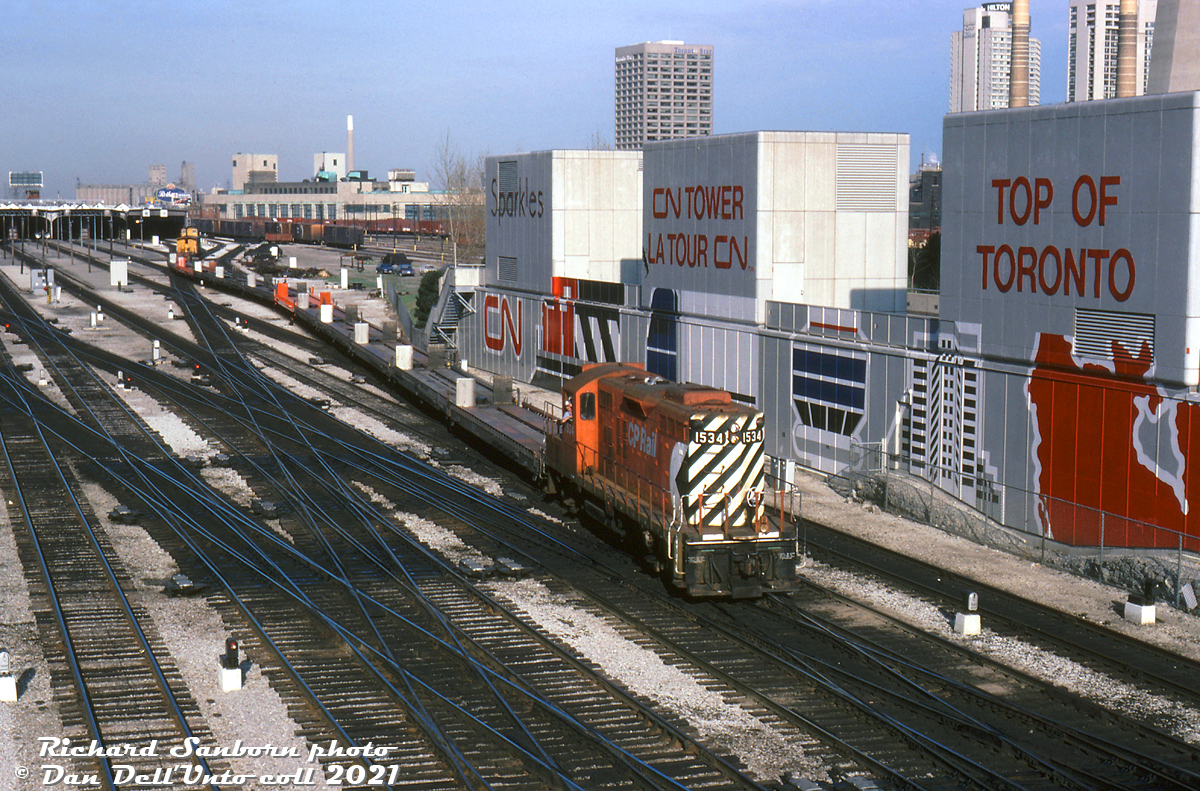The 80's in Toronto was a transitional period regarding the presence of rail activity downtown, as both CN(VIA) and CP pulled out of their respective John Street and Spadina servicing facilities as local industrial traffic dried up, and passenger servicing was consolidated to more modern facilities out of the downtown core.

CP GP9u 1534, a relatively recent rebuild done by Angus Shops just a few years earlier (ex-8538), handles a transfer with a string of piggyback flatcars and old wooden van 436994 on the tail end, heading westbound through the Toronto Terminals Railway trackage by John Street interlocking.

The base of the CN Tower is on the right with an updated CN railway mural (no more Tempo and Turbo Trains the old version had). The CP John Street yard tracks in the background that once teamed with maroon and stainless steel passenger cars now only see old boxcars, OCS (On Company Service) work equipment, and piggyback and container flatcars stored. John Street would soon be closed in 1986, and the roundhouse proper donated to the City of Toronto.

The harbourfront skyline is relatively uncluttered here: the usual buildings are present including the Victory Soya Mills elevators, the Postal Building (real estate developers Bramalea Ltd & Trizec would buy it in the early 90's, but redevelopment fell through due to the recession. It eventually became home of the Air Canada Centre), the Toronto Star office tower at 1 Yonge St., and the Hilton Harbourcastle hotel.

The two smokestacks on the right were for the nearby Central Heating Plant the provided steam heat to downtown buildings including John Street Roundhouse, Union Station, and the Royal York Hotel. It would be demolished in 1990, around the time when the land around John St. was cleared for future redevelopment. As part of the project, the former yard area was excavated and part of the old York Street viaduct under the yard was demolished, shortening the viaduct to stretching just under Union Station's train shed.

Richard Sanborn photo, Dan Dell'Unto collection slide (with thanks to Kevin Reed for passing this one on)