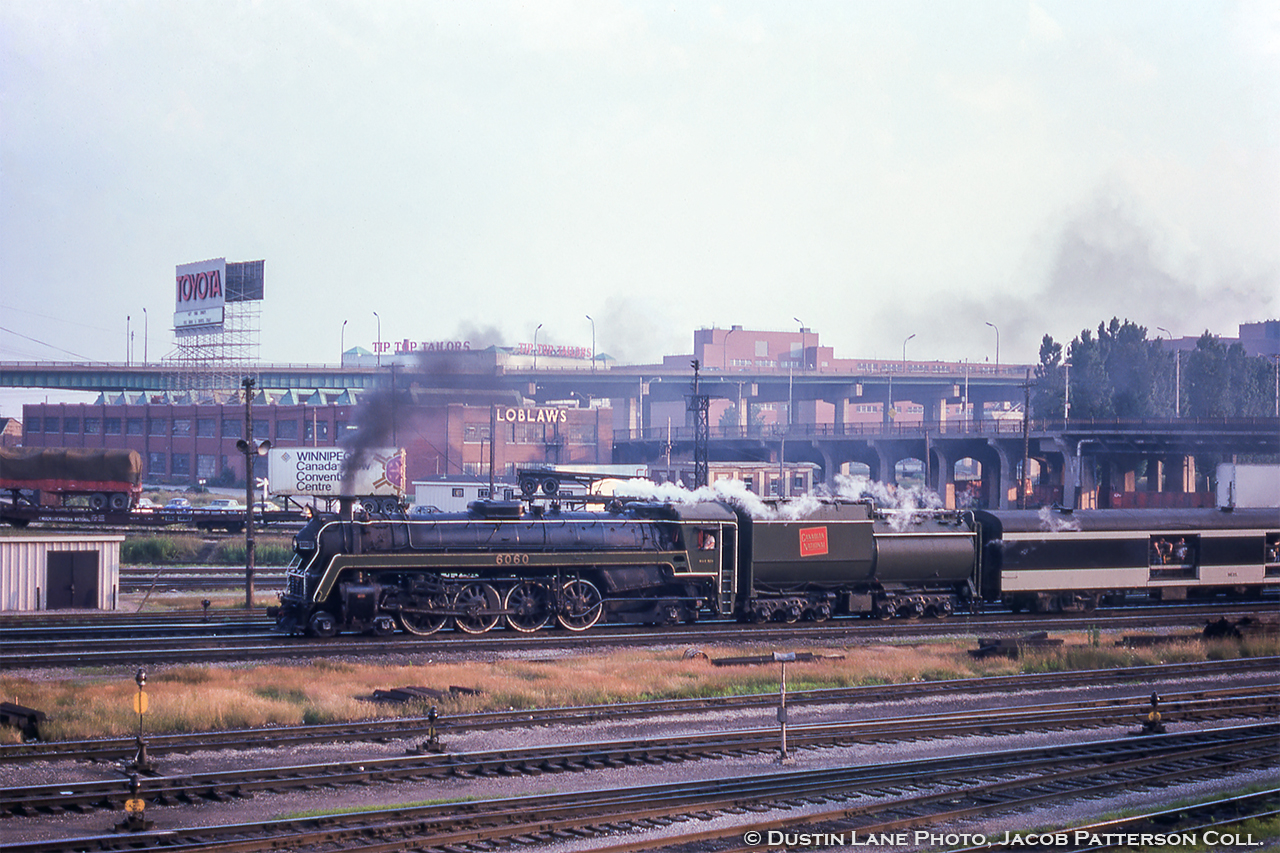 A Saturday run of CNR 6060 from Toronto to Niagara Falls and return passes beneath the Bathurst Street bridge arriving home to Toronto.  Note the signal bridge just above 6060's cab, holding the signals for the 'High Line' detour around Union Station, and the TOFC activity nearby.  One trailer advertises "WINNIPEG, Canada's New Convention Centre," most likely the Winnipeg Convention Centre, opened in 1975 as Canada's first purpose-built convention centre.  It has since been renamed the RBC Convention Centre Winnipeg in 2013.  Switch stands of the Bathurst North Yard dot the foreground, while in the background, warehouses of different sorts can be seen on Lakeshore Avenue. 

The Loblaws warehouse stands prominently just beyond the yard, and is the 1934 addition to the original 1928 office, production, and warehouse structure.  With Loblaws offices moving to St. Clair Avenue in the 1970s the building was temporarily used by the Daily Bread Food Bank during the '80s and '90s.  redevelopment plans coming in the early 2000s threatened the building, with most plans keeping the facade while demolishing the rest of the structure.  Today, the warehouse sections running north from the building along Bathurst, including the northernmost section seen above 6060, have been demolished to open space for condos, while the original 1928 office section has been restored (after being dismantled piece by piece) as the West Block.

Just above behind the plume of smoke the Tip Top Tailors plant at 637 Lakeshore Boulevard West.  Completed in 1929 to house manufacturing facilities, warehousing, offices, and retail space, the structure survives today as the Tip Top Lofts and has received a restoration of it's facade plus six floors added to the building.  Per a current (2021) listing, a 2 bedroom plus 2 bath is going for $2,499,000!

At right, the large brown set of buildings was the site of Molson's Brewery  built in 1955 and demolished in 2006.

Dustin Lane Photo, Jacob Patterson Collection Slide.