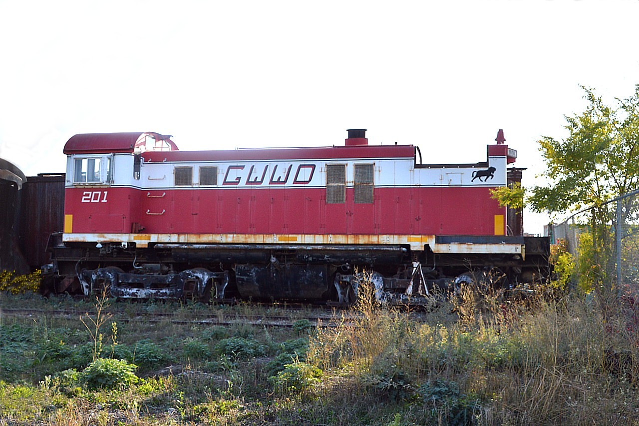 Not much more frustrating a 'treasure hunt' as wandering around Winnipeg looking for the Great Winnipeg Water District Railway. Finally, after locating it off Pinguet St in St. Boniface; we arrive only to find that it is all behind high security fencing since the City Municipal trucks and all are stored there. And worse, about 90% of the day one would not only have to deal with the tall wire topped fence, but also shooting into the sun with tall weeds to help mess up the scene.
The GWWD rarely ventures far these days; its glory days are done. The line still runs all the way down to the eastern limits of the province, some 92 miles to Indian Bay. But it is seldom used.
Only evidence of activity that could take place these days was a row of rusty GWWD lettered hoppers just outside the fencing to the south-east of the facility. 
Other than a Rail Bus and a coach, we could only see and barely photograph this old MLW S-13, built back in 1959 and looking as though it may never see the likes of freedom again.