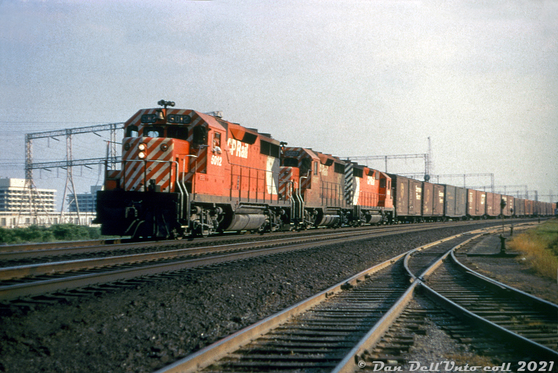 CP Rail GP35's 5012, 5007 and another 5000-series sister unit (either 5010 or 5020) roll an easbound freight along the Belleville Sub through the Leaside area of Toronto, passing by industrial leads and sidings at the east end of the yard. All three sport the new CP Rail branding, including action red paint, 5" end striping and "multimark" logos introduced two years later in 1968 (but repaints didn't generally start until mid-1969). Long an industrial center in the city, today in Leaside most if not all of the rail customers are gone, and the industrial leads and sidings have all been pulled up. What remains of Leaside Yard is a shadow of its former self.  The 2,500 horsepower GP35 model was the next iteration of EMD/GMD's mid-1960's second generation 4-axle diesel road unit after the GP30, and designed to compete with the likes of GE's U25B and Alco's C424/425 offerings. It sold quite well to US buyers, although Canadian buyers had relatively newer fleets at the time so didn't purchase all that many: CP went in for two GP30's, and three subsequent GP35 orders (that only amounted to 24 units total). Some were purchased new, others were purchased through EMD/GMD's trade-in program using old or wrecked power such as GP7's, FP7's, F7B's, etc and some components from them were reused. CN only purchased two GP35's and followed up with 15 GP40's, and later many many more GP40-2L/W units.  The GP35 model wasn't favoured by CP motive power officials, as problems cropped up with them over time and GM was reportedly resistant to working on fixes. More complex transition and wheel slip systems, more DC brushes to change in the main generators, and reliability issues (many argue the 567-block was being was being pushed to its limits by then at 2500 horsepower) didn't play in their favour, and CP de-rated them down to 2250hp at one point. Years later when they were assigned out west, a few Port Coquitlam shop employees took them under their wing and worked out many of the bugs, improving performance. Most were retired and sold off in the 90's, with a few becoming cab control units 1125-1128 (some still on the roster today) paired with GP38-2's.  Original photographer unknown, Dan Dell'Unto collection slide (duplicate)
