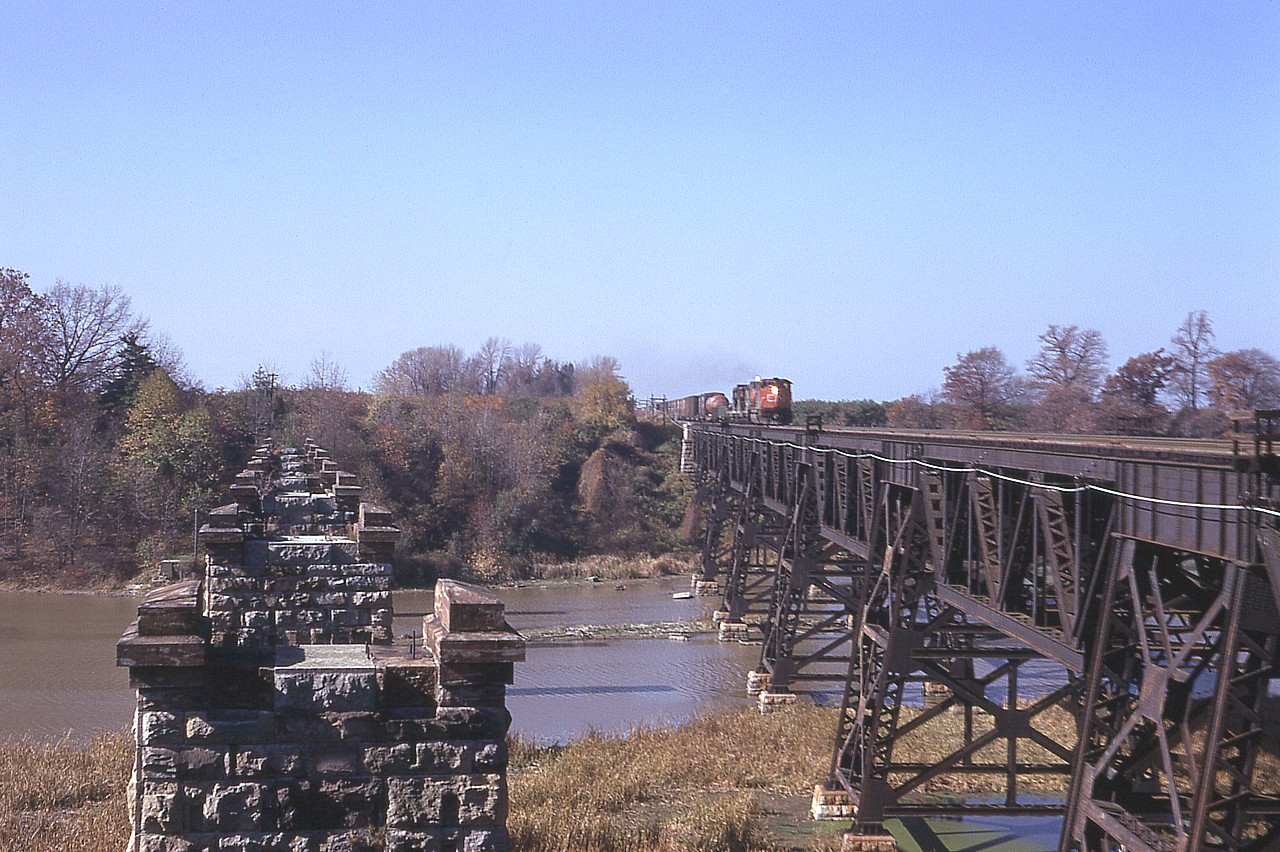 This is pretty well just an image to kindle the curiosity of those who enjoy history in Niagara. The image shows the two bridge structures over the Jordan Harbour/Twenty Mile Creek, part of the CN Grimsby sub.
It is my understanding that the first bridge, built around 1853, was wooden. But cannot verify this. (Great Western line between Hamilton and Niagara Falls opened in November 1853) The stone pillars you see on the left supported the railroad bridge built in 1867 and it remained in service until around 1900-1903; when the current bridge was built and opened for traffic. To this day it remains in use.
The structures are best viewed from down on the water, but failing that, I shot this from the east side support which is still in place, but now very difficult to access to this photo spot due to extreme foliage. There is not all that much information out there on these structures, which I find rather frustrating. Something worth looking into.  The approaching train has CN 2511 and a couple of GP9s for power, heading to Fort Erie.