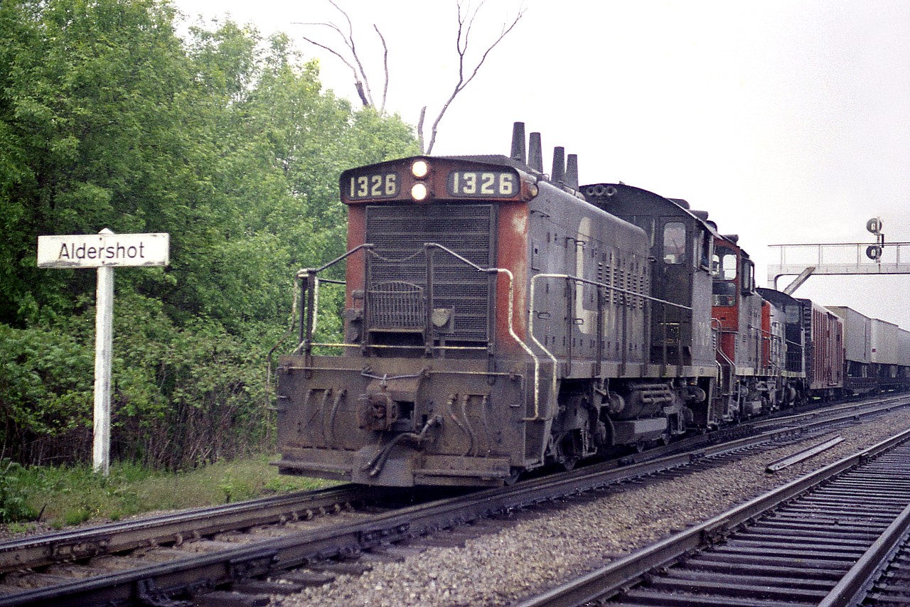 This rather mundane, I guess you could say, image has long been a favourite of mine. Why? Because it is one of the very few I managed ground level with the old wooden "Aldershot" namesign  before the area went under extensive change and became the big GO/VIA station and yard facility that it now is.
For this shot I parked over by the Aldershot Cold Storage building (now gone) on the east side of the Waterdown Road bridge, and walked eastward along the tracks. Wide open and a countryside setting, except for the Cooke Concrete block business out of sight to my right.
Forty-five years ago. And for once it actually seems that long ago.
CN 1326, 1319 and what looks like 71XX are coming off the siding with a cut of cars probably going to Hamilton.