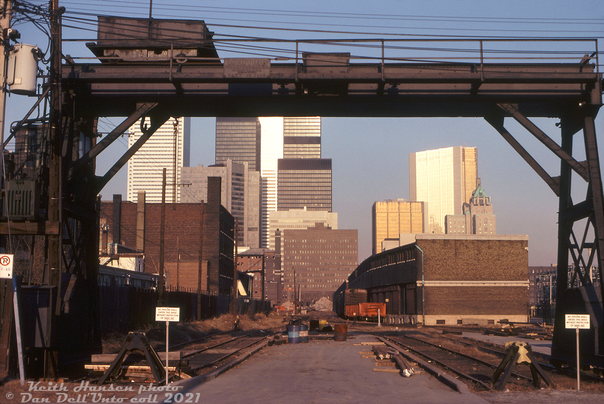 Preface: a photo exemplifying the slow decline of the railway's importance to the downtown cores of Toronto and other big cities. Once the main method to reliably transport both people and goods across Canada and into the US, the shift of manufacturing out of downtown to the suburbs and overseas, the transition from blue-collar to white-collar employment (traditional manufacturing giving way to business/finance/commerce/technology sectors of the economy, often centralized in major North American cities such as Toronto and Montreal), changing modes of transportation including the rise of truck traffic, globalization, containerization, airline travel and the private automobile meant the railways didn't really factor in as prominently in many people's lives as before, or in the operations of many of the "new" downtown businesses (other American cities were less fortunate, as historically the rise and move to the suburbs often left downtowns desolate and crime-ridden. This fate was largely avoided by Toronto and other large Canadian cities).  As such, railways followed where their customers went: sidings to new suburban business parks, new modern automated classification yards outside of the cities where land was cheap and plentiful, new piggyback and container-handling yards located near major highways and junction points, and centralized freight sorting/logistics warehouses. Demolition of many old steam-era railway facilities in and around downtowns followed, many by now either outdated, under-utilized, constrained for space, or replaced by newer facilities. CN and CP both came up with redevelopment plans for their sprawling downtown real estate holdings. The CN Tower, for instance, was just one part of a larger 70's Metro Centre development plan that never came to fruition. Even Toronto's Union Station was considered for demolition and redevelopment at one point.  --------------  Framed in the Whiting 20-ton overhead crane of the nearby team tracks, the old CP Express / Dominion Express Company freight sheds and office building at Simcoe and King Streets are slowly being reduced to rubble, in preparation for redevelopment as a new concert hall site for Massey Hall. Originally built in 1912-13, the sidings in the distance were once home to long cuts of 40' boxcars taking on and unloading LCL (Less than Carload) shipments, transloaded to/from teams of horse-drawn wagons and later trucks for local dispersal. Steam engines and early diesel switchers would regularly head up the King Street shed lead's incline from Bathurst Street, cross Spadina and head to the sheds to pull and spot cuts of boxcars, as was also the case at CN's own freight sheds located to the south (demolished around 1967-68). We're looking east from Peter Street towards the main site, bounded by King, Simcoe, Wellington and John Streets (upper-most yard in the middle here). The main CP Express offices were in a 7-story building at the SW corner of King & Simcoe (partially visible) were also to be demolished (there were some who voiced opposition to its demolition and proposed incorporating it into the new development, and the city did recognize its historic value, but ultimately it was not saved). The orange backhoe on the roof and equipment in other photos suggests local demolition contractor Teperman Wrecking is doing the work, also known for demolishing the old Eaton's Queen Street store.  New modern office towers loom high in the background, pointing to future trends to come for downtown Toronto. The new development coming here is a joint effort between CP's real estate branch Marathon Realthy, who wanted multiple office towers with commercial space, the City of Toronto (spearheaded by mayor David Crombie) who wanted to incorporate a certain amount of housing under a new planning board requirement, and the Massey Hall board of governors who wanted a site to build a new Massey Hall concert venue. Initially the project was sparked by a request from Mayor Crombie to CP CEO Ian Sinclair about using the site for a new Massey Hall. Sinclair agreed and got CP's real estate arm Marathon involved. According to a July 1976 article in the Toronto Star, after much negotiation (CP was opposed to incorporating housing in the then-still industrial area) a deal was worked out between the three parties, but some viewed the land lease terms the city agreed to as very favourable for Marathon. Maybe too favourable, as even though the plan was voted on and accepted by city council, there was some debate about it: one member quipped it was "the worst deal since Sir John A. Macdonald sold part of Canada to the railways for two pieces of steel", others considering it a "total sell-out" on the mayor's part to Marathon.  At any rate, the old freight sheds and office building were demolished, the site was cleared, and development on the new Massey Hall began. The name was changed to Roy Thomson Hall because of a donation from the Thomson family during fundraising efforts, and the new concert venue opened for the Toronto public in September of 1982.   Despite the initial agreement, other new development on the site was slow to follow, and most of the property (and adjacent former CN land to the south) remained parking lots for years until more development began in the late 80's. Notable was Metro Hall, a new office building for the pre-amalgamation Metro Toronto governing body, that would be built on the site in 1992. Some of the old CP freight sheds on the right of the photo (west of John Street) and team tracks would be retained into the mid-1980's for paper deliveries (the "Paper Shed"), but too were eventually demolished after 1987.  One small piece did manage to elude the wrecking ball: the old crossing shanty in the middle of the photo for John Street (that intersected the sidings and the sheds) would be saved, and today is preserved at the TRHA's Roundhouse Park nearby.  Keith Hansen photo, Dan Dell'Unto collection slide.