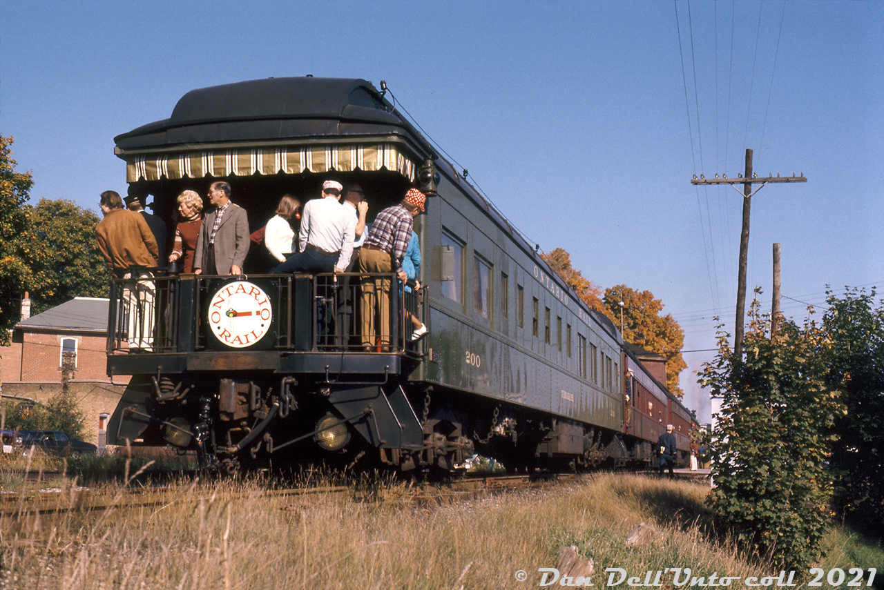 Riders crowd the rear observation platform of Ontario Rail's private car #200 "Temagami", bringing up the rear of an ORA fantrip heading up the CP Orangevville Subdivision with ex-CPR steam engines 136 & 1057 up front. The train is likely stopped to take on water at the Inglewood water tower east of the crossing, blocking both the main road through town and the CN-CP diamond (under Temagami's front truck) where CN's infrequently-used Beeton Subdivision crossed CP's Orangeville Sub (later Owen Sound Sub) in the village of Inglewood.

Temagami was originally built as business car "Ontario" for Ontario Northland predecessor Temiskaming & Northern Ontario by Pullman in 1914, later acquiring the name Temagami and number 200. She was sold to Ontario Rail Association in 1972. Today she resides at Ontario Rail's successor South Simcoe Railway as car 200, renamed "Nottawasaga".

The CN's Beeton Sub, originally part of the old Hamilton & North Western Railway from Hamilton to Barrie dating from the 1870's, was pieced up when the early 1960's "Toronto Bypass" project gobbled up the Burlington-Georgetown portion of the Milton Sub as part of the new Halton Sub, with the line north of Georgetown to Barrie becoming the Beeton Sub. One 1970's timetable notes CN wayfreights #522 and #521 were operated between Barrie and Georgetown on an "as required" basis, no doubt due to low traffic volumes. The heaviest power permitted were CN 1200-1300-series SW1200RS units. By the early 1980's a special note was added in CN Timetables that special permission was required to operate south of Beeton to the end of the line at Cheltenham. By 1984, the diamond and CN line through Inglewood had been taken up. However the Beeton-Tottenham portion would come under ownership of the Ontario Rail Association in 1986, and a name change gave them the new (and present) South Simcoe Railway, where 136, 1057 and 200 all currently reside.

Original photographer unknown, Dan Dell'Unto collection slide.

The same train near Markdale: http://www.railpictures.ca/?attachment_id=46874
On a run-by at Cataract: http://www.railpictures.ca/?attachment_id=47132