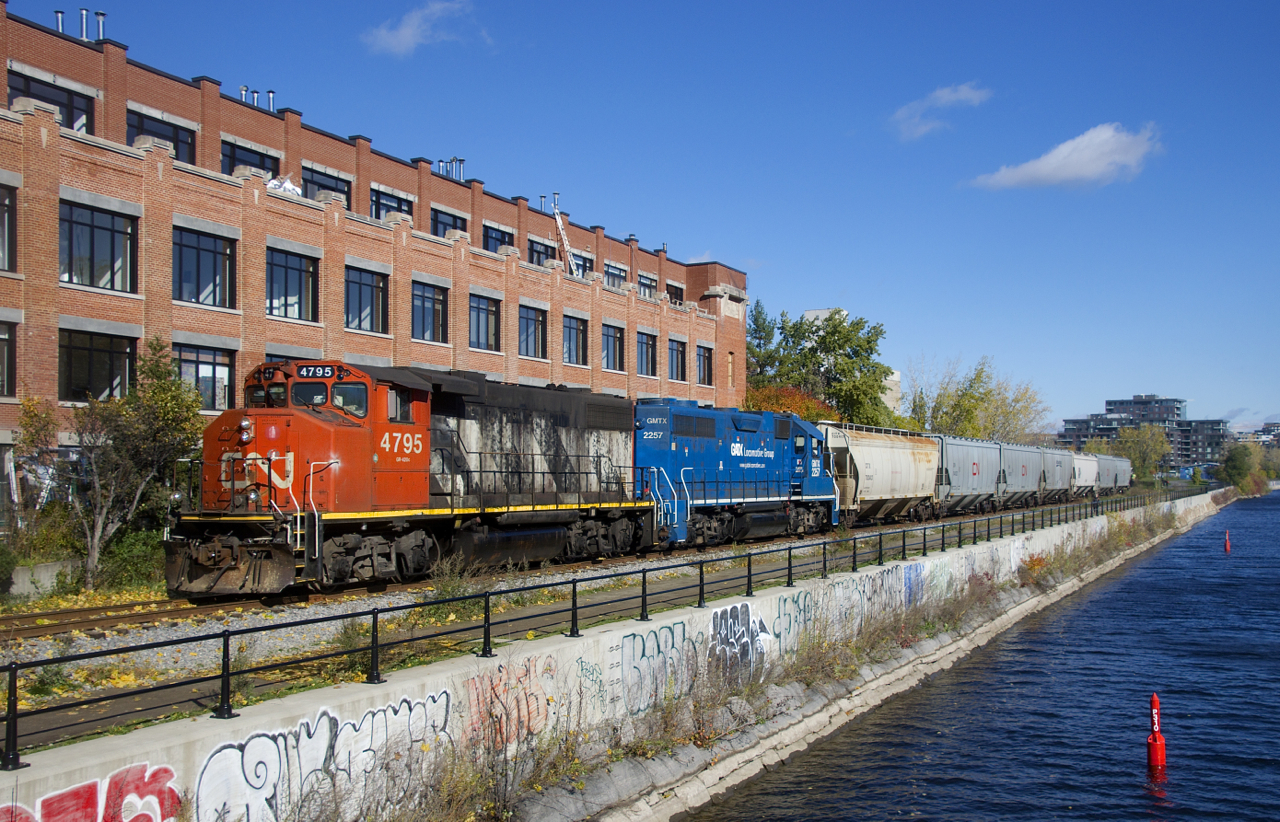 CN 4795 & GMTX 2257 are pushing seven grain loads towards Ardent Mills (mostly obscured by trees in this view) as they pass the heavily tagged retaining wall of the Lachine Canal.
