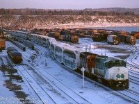 <b><i>Ready for the morning commute:</b></i> two GO Transit trainsets make their way to the <a href=http://www.railpictures.ca/?attachment_id=36825><b>CN James St. (Hamilton) Station</b></a>, deadheading out of service together through CN's Stuart Street Yard as the sun rises on a cold April morning. According to Reg's notes, the consists of today are: the first train with GO GP40-2W 708, leased <a href=http://www.railpictures.ca/?attachment_id=36391><b>CN GP40 9317</b></a> (one of two with 89mph passenger gearing) leading 10 bilevels, and APCU 900 trailing to provide the HEP. The second train coupled on has GO GP40TC's 501 and 505 sandwiching 9 bilevels.<br><br>Upon arrival at Hamilton, they will uncouple and depart on two separate morning departures eastbound for Toronto's Union Station. Timetables for that period list the pair of morning Hamilton trains as #952 (departing 6:40am) and #958 (7:15am). A morning VIA #636 (#635 from Niagara Falls, continuing as #636 to Toronto) also served the station daily at 7:40am.<br><br>Among the goodies in the yard are some barrel-style ore cars for the Dofasco ore trains, CN piggyback trailers, some tarped container loads on flats, and some blue CAST containers stacked in the distance. A CN GP9 appears to be switching at the far end of the yard.<br><br><i>Reg Button photo, Dan Dell'Unto collection slide.</i>