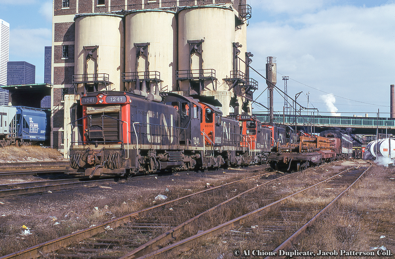 Sitting beneath the long-retired Spadina coal tower a line of GMD and MLW switchers are seen.  Note the hoppers at left to provide sand to the serving area towers, flatcars of wheel sets, and work service baggage cars.At right one of the smokestacks of the Toronto Terminal Railways' Central Heating Plant (built in 1929, twin smokestacks seen here), which supplied heat to Union Station, the Royal York Hotel, CNR and CPR express buildings, CPR John Street Roundhouse, and a number of other office buildings.  Originally burning coal, the plant was eventually converted to natural gas, and finally shutdown in the 1980s once the TTR converted to a commercial supplier, and demolished in 1990.SW1200RS 1241, built in 1956, renumbered 7300 in 1985, to CN Sweep 7100 in 1986.  MLW S4 8033, built 1954, retired in the early 80s.  SW1200Rs 1254, built 1956, retired 1993 and sold to Zyndar Bothers in Vaughan (then switching contractor at Vaughn Intermodal Terminal), renamed Plusar Inc., and finally Parsec Intermodal of Canada Ltd. 1254.  Disposition unknown.  MLW S13 8514, built 1959, rebuilt as S13u 8706 in the late '80s, retired circa 1996-97, sold to Canac, disposition unknown.More Spadina Coal Tower:Bill Thomson: 6167 being readied for an excursion; April 1963.VIA units by Pierre Fournier, April 1985.Paul Martin Photo, Jacob Patterson Collection Slide.