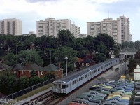 A 4-pack of Hawker Siddeley H1 subway cars are about to duck into High Park subway station, heading eastbound at Clendenan Avenue over one of the open-cut sections of the TTC's Bloor-Danforth subway line. New automobiles in a mix of early 70's colours populate the backlot of an adjacent automobile dealership along Bloor Street West.<br><br>The coming of a new east-west Toronto subway in the mid-60's spurred surrounding development along the line: the apartment buildings in the background between High Park Ave. and Keele St. sprung up around the time the new line was being built, somewhat out of place among the older detached houses of this established High Park neighhourhood. One of the casualties were the old High Park Mineral Baths, once located south of Gothic Avenue just beyond where the subway tunnel portal is in the distance.<br><br>The western end terminus of the Bloor-Danforth subway line was orignally at Keele when it opened in February 1966, but was extended to Islington in May 1968 (note rollsign on trailing car), and eventually to Kipling in November 1980 where it remains today. <br><br><i>Ted Wickson photo, Dan Dell'Unto collection slide.</i>