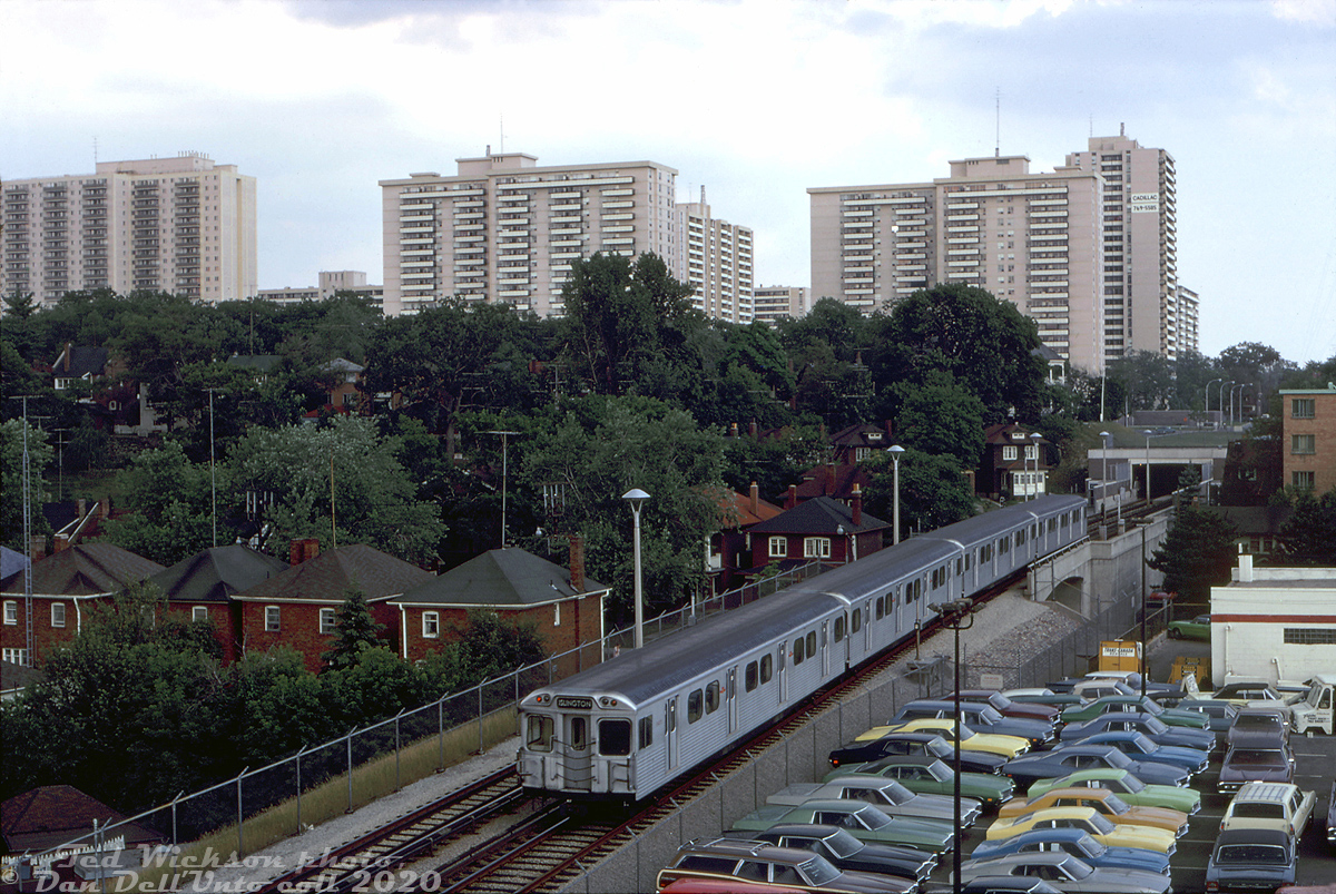 A 4-pack of Hawker Siddeley H1 subway cars are about to duck into High Park subway station, heading eastbound at Clendenan Avenue over one of the open-cut sections of the TTC's Bloor-Danforth subway line. New automobiles in a mix of early 70's colours populate the backlot of an adjacent automobile dealership along Bloor Street West.

The coming of a new east-west Toronto subway in the mid-60's spurred surrounding development along the line: the apartment buildings in the background between High Park Ave. and Keele St. sprung up around the time the new line was being built, somewhat out of place among the older detached houses of this established High Park neighhourhood. One of the casualties were the old High Park Mineral Baths, once located south of Gothic Avenue just beyond where the subway tunnel portal is in the distance.

The western end terminus of the Bloor-Danforth subway line was orignally at Keele when it opened in February 1966, but was extended to Islington in May 1968, and eventually to Kipling in November 1980 (where it remains today). 

Ted Wickson photo, Dan Dell'Unto collection slide.