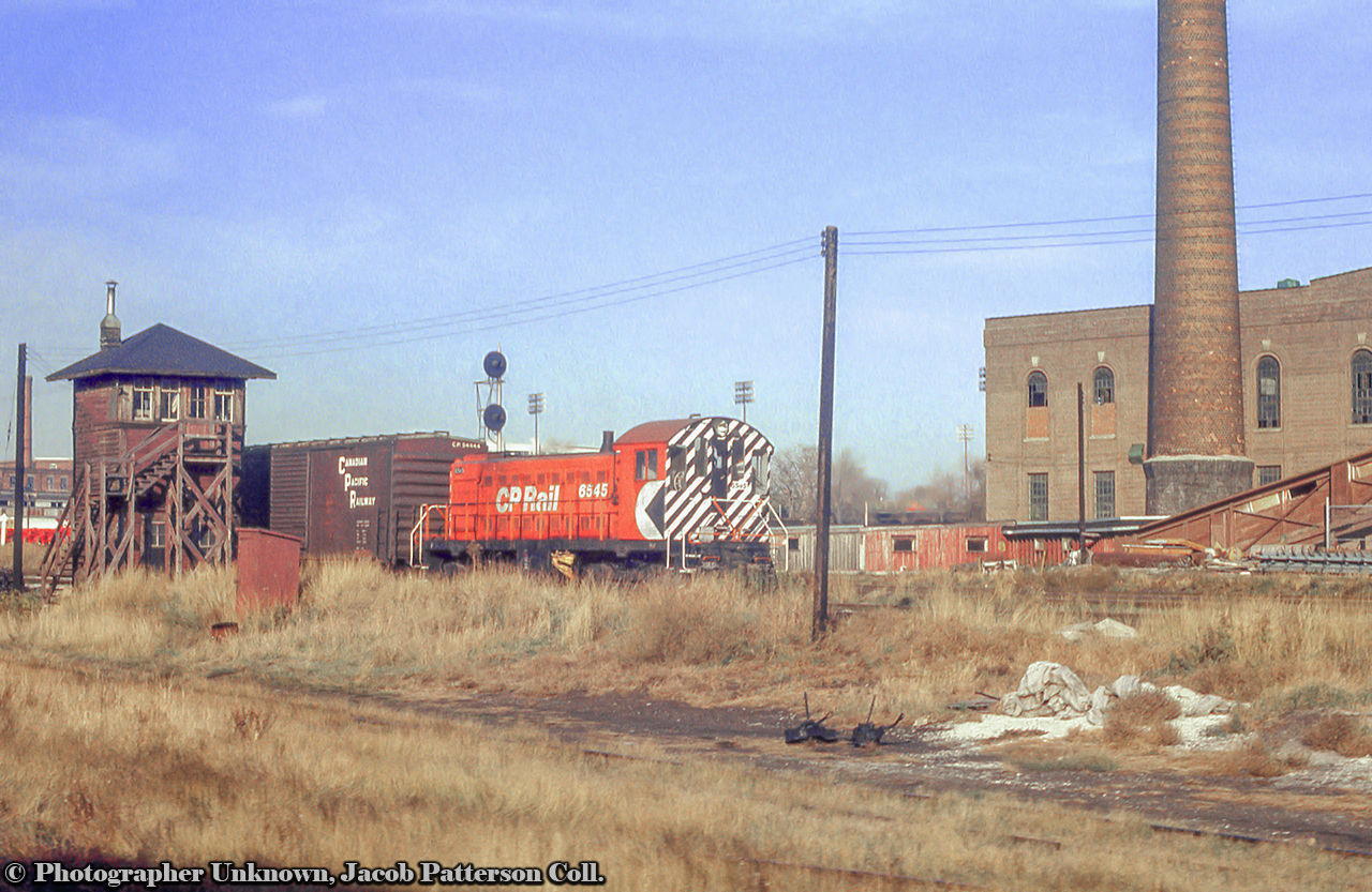 Headed downtown, CP Rail S3 6545 passes Tecumseh Street Tower with a boxcar in tow wearing the block lettering scheme.    At right the smokestack of the Wellington Destructor can be seen towering over the scene.  Originally built in 1925, the Wellington Destructor was Toronto's garbage incinerator for about fifty years, until it was closed in the mid 1970s.  At this time it was used as a transfer station until decommissioning in 1986.  Today the structure sits empty, but on Toronto's Heritage Register, and recently having undergone repairs in 2016.  In the foreground, a Toronto Terminals Railway MoW stub track sits empty with two Racor model 20B switchstands, sans targets, sitting off to the side.  CP 6545, Built by MLW in 1955, will serve the CP until retirement and scrapping in 1986.6545 at West Toronto.Original Photographer Unknown, Jacob Patterson Collection Slide.