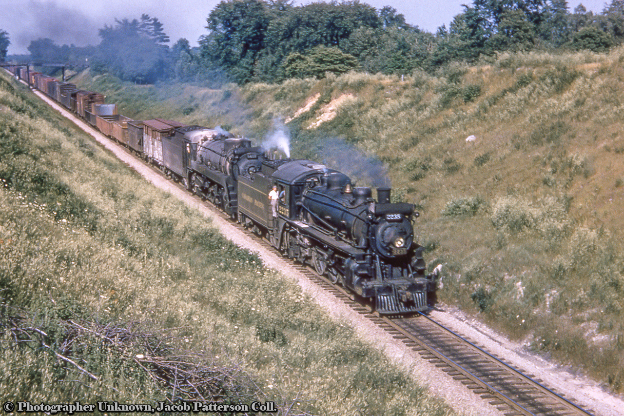 A westbound freight behind heavy Mikado 5417 with Pacific 2235 on the point as a helper approaches the wooden bridge just east of Galt.  The helper will stay on until cresting the grade at Orr's Lake, where it will duck in the siding, coast back down to Galt, head around the wye, and run light back to Toronto.  Note the brakeman hanging out the door.  There was once two farm bridges at this location, both of which can be seen in this 1945 aerial.Original Photographer Unknown, Jacob Patterson Collection Slide.