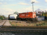 CP RS18 8751, handling today's "Moonlight", is captured here in a rare photo working the CN-CP interchange at Brampton diamond during its trip south on the Orangeville Sub enroute to Streetsville Junction. The unit has uncoupled from its train (visible sitting on the main track in the distance) and is backing around on the south leg of the interchange track, either lifting or setting off cars. Note the neat NCHX covered hopper lettered for CIL (Canadian Industries Ltd) just behind the power. The diamonds for the CN Halton Sub north and south mainline tracks are visible at the lower left. <br><br> 1950's maps show the Brampton interchange track split off from the Orangeville Sub just south of Rosedale Ave. and continued south to the diamond, where it curved east around the south end of the J.W. Hewetson Shoe Co. building, crossed Mill Street and lead into the CN station grounds. By time this photo was taken, the interchange had been extended north to just south of English Street, and at the south end the track ended to the right just out of frame at a switch to CN's Halton Sub (protected by dwarf signals), that CN used to access the interchange. One can see a crossover north of the diamond that allowed CP to come off the Orangeville Sub and access the south end of the interchange track for southbound lifts and setoffs (best reference online I can find is this <a href=http://jpeg2000.eloquent-systems.com/toronto.html?image=ser12/s0012_fl1968_it0289.jp2><b>1968 aerial</b></a>). <br><br> North of the diamond just south of Vodden was a siding that branched off the interchange track and headed east to Dale Estates (a large Brampton greenhouse operation, helping give Brampton its "Flower City" nickname). There are accounts of fresh flowers being shipped out of the CPR Brampton Station on passenger trains back in the day, but apparently the boilerhouse at the end of the spur once took hoppers full of coal. <br><br> I haven't been able to nail down an exact date when interchanging here between CN & CP was discontinued, but have heard it was sometime in the 80's. A source mentioned some modern OBRY traffic for CN was still billed as interchanged at Brampton (on paper), even though the physical interchange actually happened at Streetsville with CP, who then forwarded the cars to CN. <br><br> The mothballed Brampton interchange track remained mostly intact and connected to CN into the 2000's until signal upgrades and track work in the area around June 2007. It was during that time when CN removed their switch and dwarfs for the interchange track, and some of the rail at the sound end was buried (the northbound and southbound OBRY signals were also replaced, as CN maintained the signal system while CP maintained the physical diamond, even in the OBRY era). Some speculate OBRY having an active interchange here with CN would have allowed them to reduce their operating costs by abandoning or selling off the southern portion of their ex-CP Orangeville/Owen Sound Sub between Brampton diamond and Streetsville, but nothing ever became of that idea. <br><br> <i>Roger Heed photo, Dan Dell'Unto collection slide.</i>