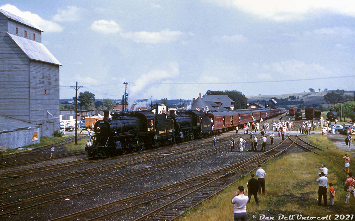 Canadian Pacific Railway D10h's 1098, 1092, and their passenger consist are seen at Orangeville Yard, after arriving northbound on a Buffalo NRHS chapter fantrip operating northbound from Streetsville to Orangeville. Riders are out and about crowding the yard near Town Line crossing, that intersected the north end of the yard. Some have climbed on the tenders of the two D10's, possibly to watch as they take on water at the adjacent water spout.

The top of the original CPR station (since moved and today a restaurant) is visible above the baggage car, and behind that the bunkhouse (burned down in 2006), freight sheds further back, and angled coal tower in the distance near the roundhouse. There were tracks behind the station that lead to coal and oil dealers, grain elevators, the stock pens, all local amenities for a small railway town. In the early 1900's, the original engine house, turntable and coaling platforms used to be located north of the crossing where this photo was taken from, but were relocated to the south at some point in time. One can see part of the Niagara Escarpment rising up in the distance.

What's really interesting about this photo is, judging by some of the people in the shot, it was taken at the same time as this photo by Bill Thomson. One of the figures at the lower right could very well be a young Bill taking his photo, captured by this unknown photographer standing on top of some freight cars nearby.

Original photographer unknown, Dan Dell'Unto collection slide.

More photos of this fantrip:
Streetsville: http://www.railpictures.ca/?attachment_id=33872
Cataract: http://www.railpictures.ca/?attachment_id=28108
Charleston Sideroad: http://www.railpictures.ca/?attachment_id=36987