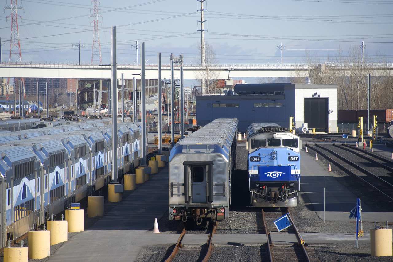 Multilevel cars as well as stored MR-90 cars are visible at the Pointe-Saint-Charles Maintenance Centre, with AMT 1347 at the front of a Mascouche trainset at right. Barely visible at left is EXO 808, heading towards the Victoria Bridge.