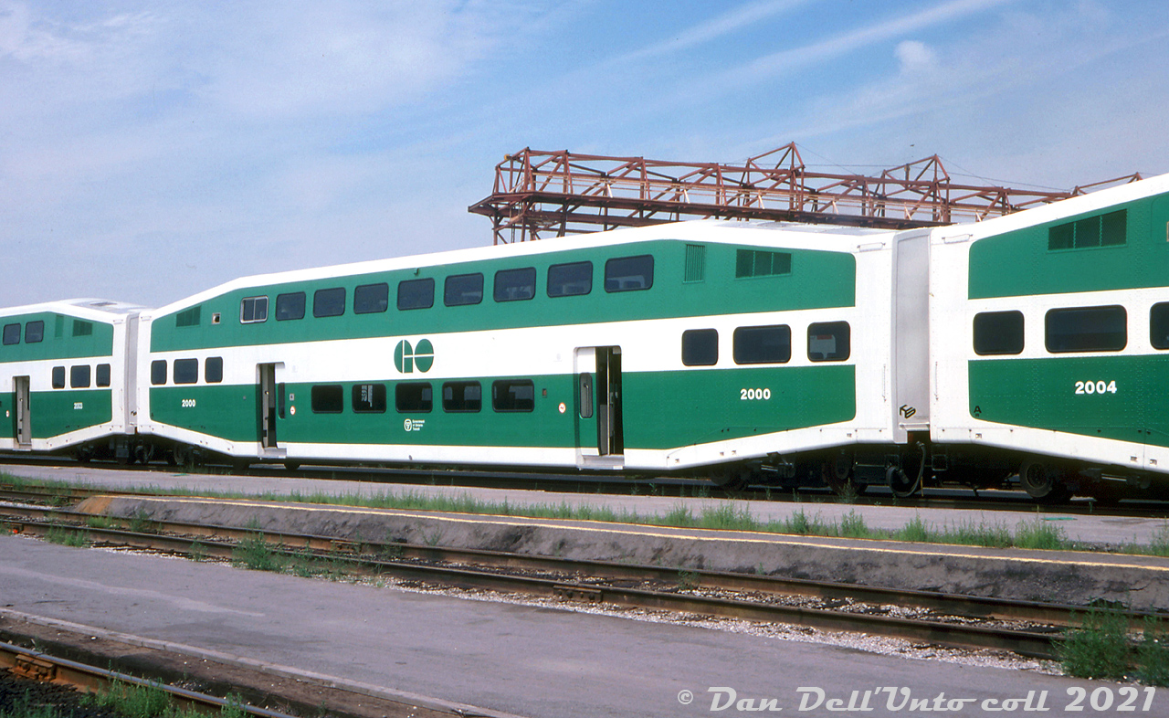 The big kahuna, the original, numero uno: the first car. GO Transit 2000, the first Bilevel commuter car built, sits in Willowbrook Yard adjacent to the canopy (that was under construction at the time). Note the neat stylized "HS" Hawker Siddeley Canada block logos on the ends of the cars.Looking for a higher-capacity alternative to its single level cars, and faced with the decision of running longer trains (and adjusting infrastructure to match) or higher capacity cars, in the 1970's GO Transit experimented with a few different commuter car designs, including borrowing both the C&NW gallery cars, and the CP ones built by Canadian Vickers. Not satisfied with the gallery car design, GO worked with Hawker Siddeley to make their own double deck commuter car design, dubbed the "Bilevel" commuter car, with two full levels in the middle and intermediate levels at each end, and two sets of doors to improve passenger flow.The initial 1977 order was for 80 coach cars (2000-2079), and over four decades later GO has amassed a fleet of over 900 bilevels, including most of the originals. Subsequent orders included cab cars, and "accessible" cars with lower door controls (the initial conductors controls were located by the silver-framed upper sliding windows) and wheelchair ramps. The design changed hands over time, being first built by Hawker Siddeley Canada, then UTDC/Can-Car, Bombardier, and today Alstom Transport (all in the old Can-Car Thunder Bay, Ontario plant). Other commuter agencies also embraced the bilevel over time, and in recent years the design has had an upgrade with crumple zones to improve crash-worthiness, and a new cab car end design.GO 2000 was one of 16 cars sold to Texas commuter startup Trinity Rail Express in 1997 (along with F59PH's 565-568), and became TRWX car 1050. The lowest-numbered bilevel on GO's roster today is 2002.Original photographer unknown, Dan Dell'Unto collection slide.