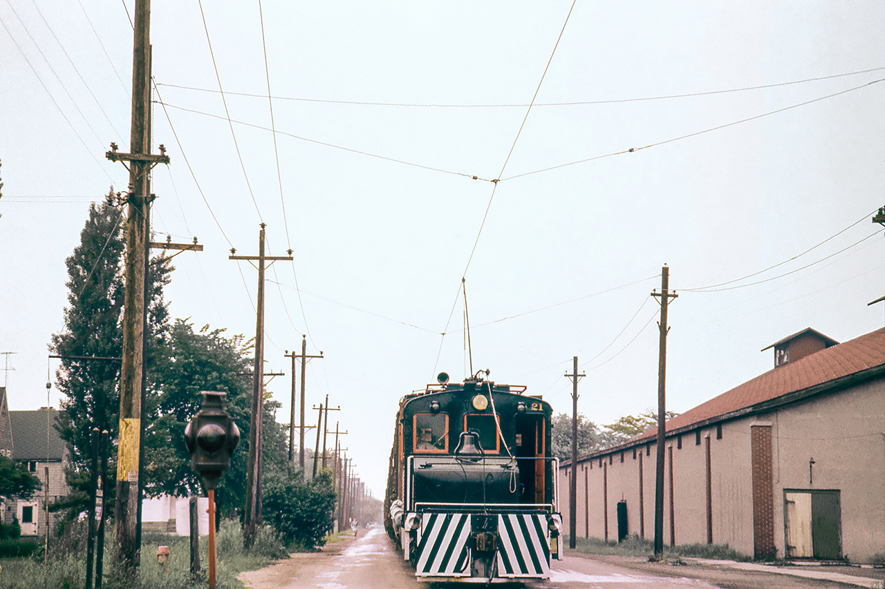 *** Submitted with the help of JACOB PATTERSON - Thank-you MIKE ***


The click of a shutter... and what do we have here... NS&T Electric Motor #21 travelling south on Elm Street approaching the Killaly Street intersection.  The train would soon pass the non-interlocked (flagged) industrial diamond for the Canada Cement company spur that connected the factory to loading docks on the Welland Canal.  The final destination for the train would be the Macey Yard Interchange with the CNR.  The train is hauling boxcars from Robin Hood Flour Mills.  The NS&T was the last railway to reach Port Colborne in 1911, and the construction of Robin Hood in 1940 created a significant source of Freight Revenue for the Company, enough to dedicate an Electric Motor to servicing the facility.  NS&T generally kept Motor #20 or 21 stationed adjacent the current Museum grounds.  #21 was built by the CLC - Canada Locomotive Company in 1927 for the Montreal & Southern Counties coming over to NS&T in 1941, likely due to freight demands at Robin Hood.  The building on the right was the privately built Port Colborne Athletic Club Arena (also called the White Arena), a 2200 seat hockey rink constructed in 1932 that launched the NHL career of of 5 time Stanley Cup Champion- Toronto Maple Leafs Captain - Ted "Teeder" Kennedy who joined the Leafs at age 16.  According to the NS&T author John Mills the last Interurban Trolley passenger run in North America was Thorold to Port Colborne - March 1959.  Electric Motor freight operations continued until the summer of 1960 at which time the line was serviced by Diesel locomotives.  Street running to the Robin Hood terminated in 1983 (or '84) when the tracks were removed as the line was re-routed from Welland south to Robin Hood and the then new "Canada Starch" plant.  The scene looks very much different today, the Cape Cod style homes on the left are still there, the rink was demolished in the late '60's, streets widened and gentrified.