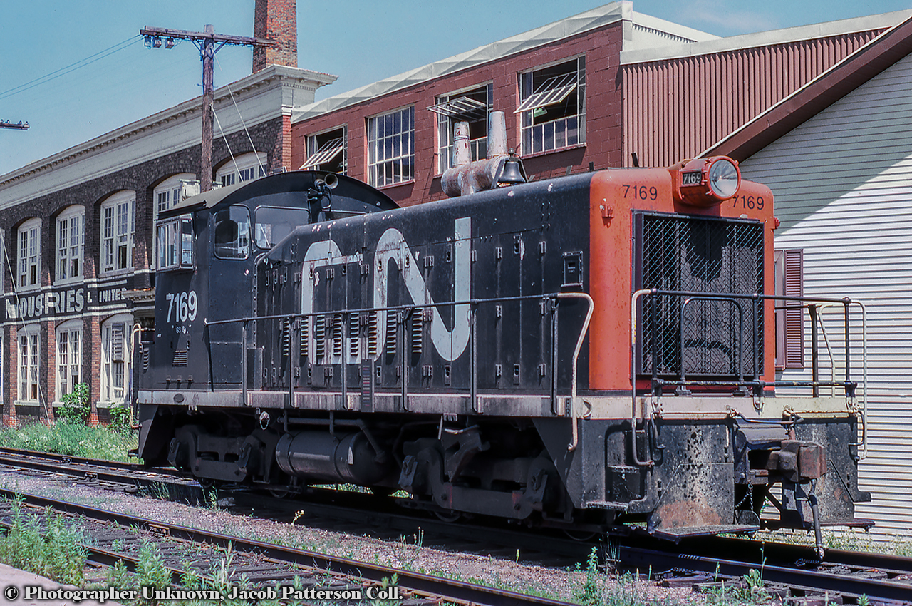 The CN Guelph yard power is seen parked in track XW41, the southernmost track across from CN's Guelph station.  Power is CN 7169, an 800hp GMD SW8 built in 1951, originally one of the 34 SW8s numbered in the 8500 - 8533 series prior to 1956 renumbering to 7150 - 7183.  All would be off the roster by 1989.Behind the unit sits two buildings, an older wood and timber structure at right at 72 Farquhar Street, and the brick factory on the left at 76 Farquhar Street, both part of Guelph's textile and agriculture history.  The "medium" structure at centre was an addition during the 1920s to connect the two main buildings.  At right, Guelph's Drill Hall built 1866 served the military for practicing various drills until the 1890s.  The armoury would not be built until 1906, directly across the street and still in use today.  The site first became part of Guelph's textile manufacturing sector in the early 1890s as part of the shirt making company, Williams, Green & Rome, per an 1892 fire insurance map.  By the early 1900s the factory at left had been built and the full site was in use as the Guelph Cotton Mills per a 1907 fire insurance map, and by 1908, taken over by the Louden Machinery Company (the word "Louden" barely visible in this 1908 photo) for the manufacturing of agricultural equipment.  The Aspinwall Manufacturing Company would assume operation of the site in 1912 as their only Canadian plant (seen here circa 1915) making equipment for potato farmers, including their automated potato planters, sprayers, cutters, sorters, and weeders.  Some examples of their equipment seen here.  Per a 1929 fire insurance map, the properties were once again in the textile industry under the Regent Knitting Company, and later by Zephyr Looms and Textiles Inc. in the late 1930s.  Zephyr Looms & Textiles worked through the Second World War handling military contracts for uniform articles and other small items.  Emerging from the war, the company would rebrand as Textile Industries Ltd. in December 1945, which it would operate as until closing for good in December of 1980.  At some point during the 1970s a slight change in the name recognized their location in Guelph on Wyndham Street, as they became known as Wyndham Textile Industries Limited.Original Photographer Unknown, Jacob Patterson Collection Slide.