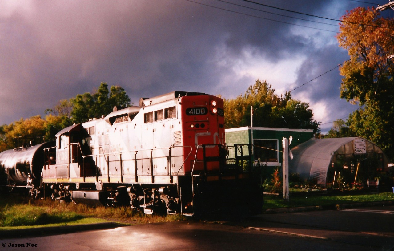 During a rain filled late summer evening I had heard the CN 15:30 Job returning through Waterloo not far from my house near the Waterloo Spur. I grabbed my Kodak camera and hopped on my bike heading to the tracks where they crossed my street hoping for the weather to let up enough for me to get a photo. As I followed the spur the rain eventually slowed to a trickle and stopped as I neared the Roger Street crossing. As the train was gaining on me, I decided this was going to be as good as it was going to get and then just like young railfan luck, the dark swirling clouds parted for a brief minute with even the sun managing to make an unexpected appearance. Here, CN GP9RM 4108 is pictured returning from Elmira to the Kitchener yard with one rain soaked tanker crossing Roger Street beside the one thriving nursery and greenhouse located beside the spur.