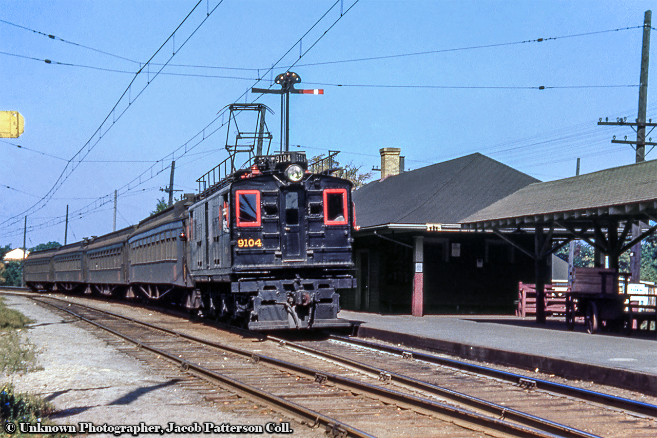 Canadian National Railway 9104 begins its journey at Saint Eustache sur le lac with train 360 and a consist made up of 4 wooden commuter coaches.  Note the truss rods, the tilted 'wafer' logo on the side of the cars, the sagging on the first car, and the two sets of marker lanterns, allowing the motor to run around the train without needing to relocate lanterns.  Scheduled 7 days per week, as per the 1948 timetable, 360 will depart at 1330h inbound to Montreal.The necessity of these locomotives ties directly to the geography of the line.  In the early 1910s the Canadian Northern Railway was in need of a direct route into downtown Montreal, aiming to avoid both the competing Grand Trunk and Canadian Pacific Railways in the process.  With access from the east, west, and south limited, the railway chose to approach from the north by boring a tunnel beneath Mount Royal.  Coming in at approximately 3.1 miles in length, the tunnel would be unsuitable for steam locomotives due to the fumes and smoke, leading to the CNoR looking to GE for electric locomotives.Built by General Electric from 1914 through 1917, the six box cab locomotives, numbered 600 - 605, were geared for 55mph passenger service under the 2,400 volt DC system running from downtown Montreal to Val Royal station, opening for service October 21, 1918.  The electrification would later be extended to Saint Eustache sur le lac in the late 1920s (see 1945 view of station here).  With the nationalization of numerous Canadian railways under the Canadian National Railways banner beginning in 1919, the locomotives would be renumbered to CNR 9100 - 9105 that year.  They would retain these numbers until November 1949, when they would lose their 9's, becoming 100 - 105.  In February 1969, they would be renumbered a final time to 6710 - 6715.With the growth of transit in and around Montreal, commuter operations had been taken over by the Commission de Transport de la Communauté Urbaine de Montréal (CTCUM - later STCUM) in 1982, with CN providing equipment, crews, and maintenance.  The end for such aged equipment was to come in the mid 1990s as new MR90 cars were ordered from Bombardier.  Retirement of these and other electric motors serving these lines came on June 2, 1995 as equipment and infrastructure were upgraded, including a new 25kv electrical system, and CTC installation, eliminating the few remaining sections of train order operation in Canada.  Agence Métropolitaine de Transport/AMT would fully take over operations on January 1, 1996.  Of note, all GE box cabs retained their wooden doors and windows, as seen above on 9104, almost until retirement.  They would be upgraded to steel in 1992 (see 6711 photo below).Of the original six, four box cabs have been preserved: 6710 at Deux-Montagnes station(1), 6711 at Exporail(2), 6714, former 9104, at the Connecticut Trolley Museum, East Windsor, Connecticut(3), and 6715 at the Museum of Science & Technology in Ottawa(4).It is worth noting CN had other electric power for these commuter operations including Electric Multiple Unit cars which will be detailed in a future post.  As for locomotives, CN also rostered three steeple cab units built by GE in 1950, numbered 200 - 202, later 6725 - 6727, and another set of box cabs - nine in total - built by English Electric in Preston, Lancashire, England.  These had been built for the Port of Montreal in 1924 - 1926 as the (now old) Port aimed to reduce smoke and fumes present in the area.  Like the Mount Royal Tunnel a few years earlier, they too ran a 2,400 volt DC system.  Numbered 101 - 109, the Commissaires du Port de Montreal motors served until 1940 when electric operations were discontinued at the Port due to excessive costs.  The motors were exchanged to the CNR, who at the time was looking to increase its roster of electric power for commuter operations.  In return, the CNR exchanged handful of 0-6-0 switchers to the Port.  Further reading on Port of Montreal electrification can be found here.CN GE box cabs:Through East Junction.CN 6714 (9104) in the noodle scheme.CN English Electric box cabs:At Val Royal.Side Profile at Pointe St. Charles.CN GE Steeple cabs:Light power at East Junction.Double header at Val Royal.Information per a number of UCRS and CRHA newsletters.Original Photographer Unknown, Jacob Patterson Collection Slide Duplicate.