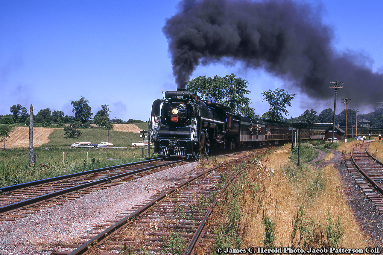Running out its final miles, Canadian National Railway 6218 rolls an excursion train eastbound through Madoc Junction on the Campbellford Subdivision returning to Belleville after turning on the wye at Anson Junction.  Having just crossed Tuftsville Road, the train passes the little station and the siding (designation DB32) in the foreground.  Note the yellow, wooden derail post.  At right lies the Madoc Subdivision.  This excursion would be one of five trips out to Anson Junction and back on July 3 with two trips to follow on July 4 to celebrate the retirement of 6218.CN’s Campbellford Subdivision was constructed in segments by different railways over almost three decades.  The earliest portion of the 86.4 mile long line was constructed by the Port Hope, Lindsay & Beaverton Railway from Omemee north west to Lindsay in 1857.  The Grand Junction Railway, originally chartered to build from Belleville to Toronto by looping north through many small communities, was altered to run Belleville to Peterborough with future expansion to Georgian Bay – later cut back to Lindsay.  Only the section to Peterborough would be built under this railway.  Construction began during the 1870s and was completed after many financial issues in 1880.  The final piece of the Campbellford Sub missing at this time was the 13 mile stretch from Peterborough to Omemee, which would be constructed by the Midland Railway of Canada in 1883.  At this time, the PHL&B Ry had been renamed the Midland Railway in 1869, though the company would grow to over four times its size in 1882 with the merger of six struggling railways under its name.  These would include the Midland Railway of Canada; Grand Junction Railway (of Canada); Belleville & North Hastings Railway (see below); Victoria Railway; Whitby, Port Perry & Lindsay Railway; and the Toronto & Nipissing Railway.  The expanded Midland Railway would be leased by the Grand Trunk Railway on January 1, 1884, and finally purchased on April 1, 1893.  The line continued service under the Canadian National Railway banner until abandonment in pieces beginning in 1987 by abandoning Corbyville (mile 3.2) and Peterborough (mile 60.6), and finally the section from Peterborough to Lindsay (mile 86.4) in 1989.Originally constructed in 1879 by the Belleville and North Hastings Railway from Madoc Junction (mile 0) to Madoc (mile 14.9), the line was leased to the Grand Junction Railway soon after completion, finally coming under control of the Midland Railway of Canada in 1881.  The Midland Railway extended the line from Madoc to Eldorado at mile 21.69 in 1887, though would close this portion in 1889 due to low traffic.  Service continued after the Midland’s merger with the Grand Trunk and later Canadian National until its last revenue movements in 1979 (5 inbound loads, 12 outbound loads at Madoc) and the line closed to traffic effective January 1980.  CN would be paying to truck loads to/from team tracks at Foxboro (mile 6.54 Campbellford Sub – image below) and Belleville.For the two days of July 3rd and 4th, 1971, Belleville was crowded with railfans from all over, many from the USA familiar with 6218 trips on Central Vermont and Grand Trunk Western lines.  A ceremony was held at Belleville station on the 4th bidding farewell to 6218 before its final departure, well documented in the Upper Canada Railway Society’s July 1971 newsletter (link below).An excerpt from the UCRS article:“6218 herself was in pristine condition for her final hours of glory.  She had been restored to her World War II appearance, complete with rectangular herald on the tender coal bunker, smoke deflectors (or "elephant ears") around the smokebox, a glossy black paint job complete with white striping on the running boards and cab of the engine and on the tender, and yellow numerals on the cab sides.  The wheels and drivers on engine and tender were painted their usual white...The highways and byways from Belleville to Anson Junction were dotted with cars with many United States license plates, as people came from far and near to see 6218 make her last run on this weekend...A stirring eulogy to 6218 was given by Mr. O.S.A. Lavallee of Montreal.  At the conclusion of the eulogy, the Duke of Edinburgh Pipe Band played "Auld Lange Syne".  There was now a hush in the crowd, as people stood waiting, cameras and tape recorders at the ready.  The band played a second time.  After what seemed an eternity, out of the east a plume of black smoke arose.  6218 then came screaming down the yard, through the leads and past the station and the assembled crowd, whistle blowing, doing an estimated 60 per.  She disappeared into the sun in the west, and it was all over.”The full UCRS article covering the final trips of 6218 can be FOUND HERE in newsletter 306: July 1971.More Campbellford/Madoc Sub images:Lindsay, Bruce Lowe, June 1964Foxboro, Doug Page, June 1964Madoc Junction, Arnold Mooney, July 1976Madoc, Arnold Mooney, July 1976Hastings, Steve Danko, May 1977Peterborough, Eric May, 1979Stirling, Arnold Mooney, July 1979Stirling, Arnold Mooney, July 1979James C. Herold Photo,  Jacob Patterson Collection Slide.