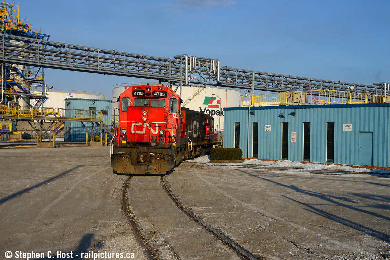 The Hamilton 1500 yard job with CN 4705 and a GP9 is heading into Bunge's parking lot, crossing their industrial lead. The gp9 is crossing over the diamond in this picture, with that track also buried in the pavement. The crew are heading to switch Vopak in the background on this beautifully sunny Winter day.