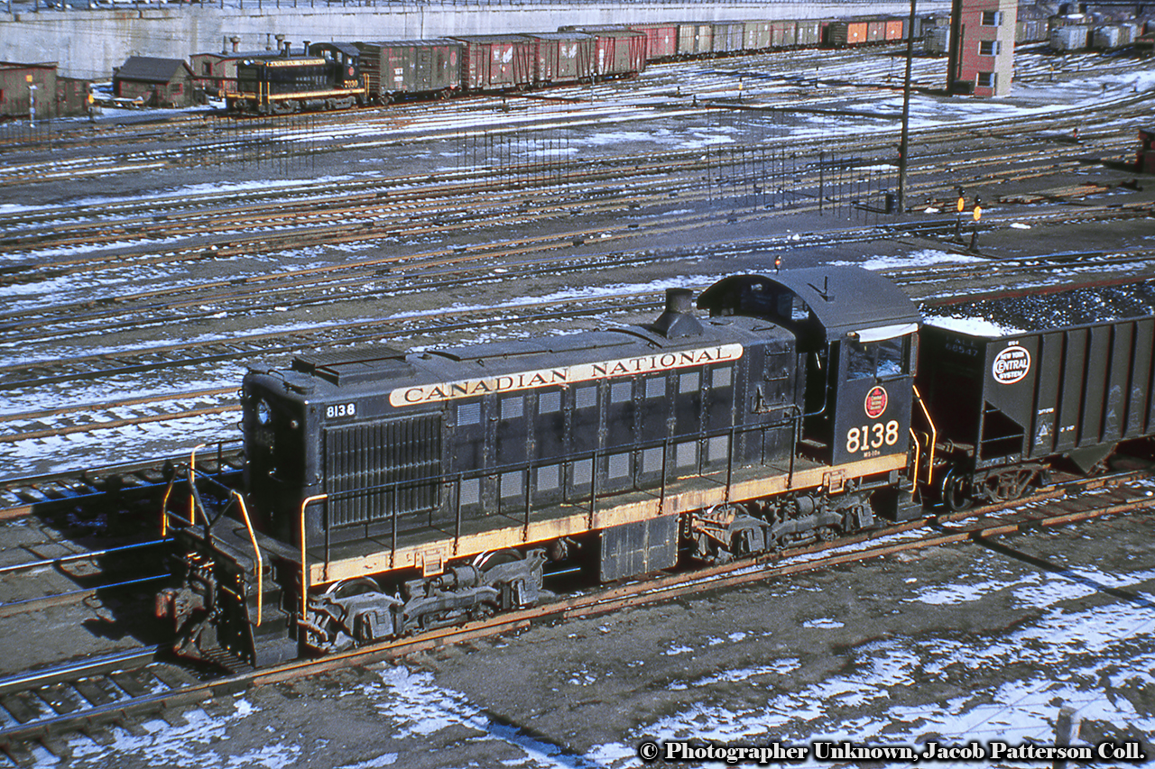 A pair of first generation MLW and GMD switchers shuffle cars around downtown Toronto at Bathurst Street, note the telltails for the bridge.  8138 is seen moving a hopper of coal around the yard leads, perhaps destined for the nearby coaling tower at CN Spadina, while 7030 is moving boxcars about Bathurst Yard.  Note the relatively new yard tower at upper right.  Built in 1949, and originally numbered 7991, the MLW S2 would be renumbered 8138 in 1956.  All of these 1000hp units would be retired by 1975.  The SW1200 7030 in the background was built in 1956 by GMD London, and would be renumbered 7730 in 1985.  She would be retired in August 1990 and sent for scrapping.Original Photographer Unknown, Jacob Patterson Collection Slide.