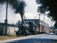 Returning from Tilsonburg with a string of mostly 40 foot boxcars, Canadian National Railway E-10-a, 2-6-0 Mogul, 92 rolls alongside Clarence Street in Brantford, just about to cross Nelson Street.  Behind the train is the large H. E Mott Company Limited building on the north east corner of Clarence and Wellington Streets, a site connected to Brantford's agricultural heritage.<br><br>Originally built in the 1870s for the J. O. Wisner Son & Co. (founded 1872) manufacturing fanning mills and seed drills, the company amalgamated with the Massey-Harris Company in 1892.  At that time, the Verity Plow Company, originally of Exeter, Ontario, relocated to Brantford in the former Wisner facility.  Verity too would become connected to Massey-Harris during the late 1890s, and the building would be sold to the Goold, Shapley & Muir Co. who manufactured farming implements with a focus on gasoline engines, used mainly in powering tractors.  Facing financial difficulties during the 1930s, the company finally succumbed to the Great Depression in 1934, at which time, the H. E. Mott Company Limited was founded, assuming control of the property until 1958, when the company relocated to a spot on Wadsworth Street.  Further information is not noted, however the building was demolished on August 31, 1976.  Information from Brantford Historian, Jack Jackowetz.<br><br>Built by the Canadian Locomotive Company in 1910 for the Grand Trunk Railway as E12 class number 1017.  It would be renumbered to CNR 919 in 1923, and to CNR 92 in 1951.  92 is one of the surviving CNR Moguls in the United States, currently awaiting restoration at the <a href=https://www.railpictures.net/photo/105947/>Wilmington & Western Railroad</a> in Wilmington, Delaware.<br><br><i>Original Photographer Unknown, Jacob Patterson Collection Slide.</i>