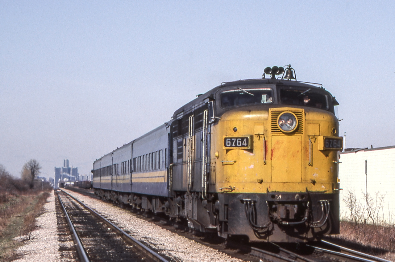VIA 6764 is eastbound in Tecumseh, Ontario on March 29, 1983. That is the skyline of Detroit in the background.