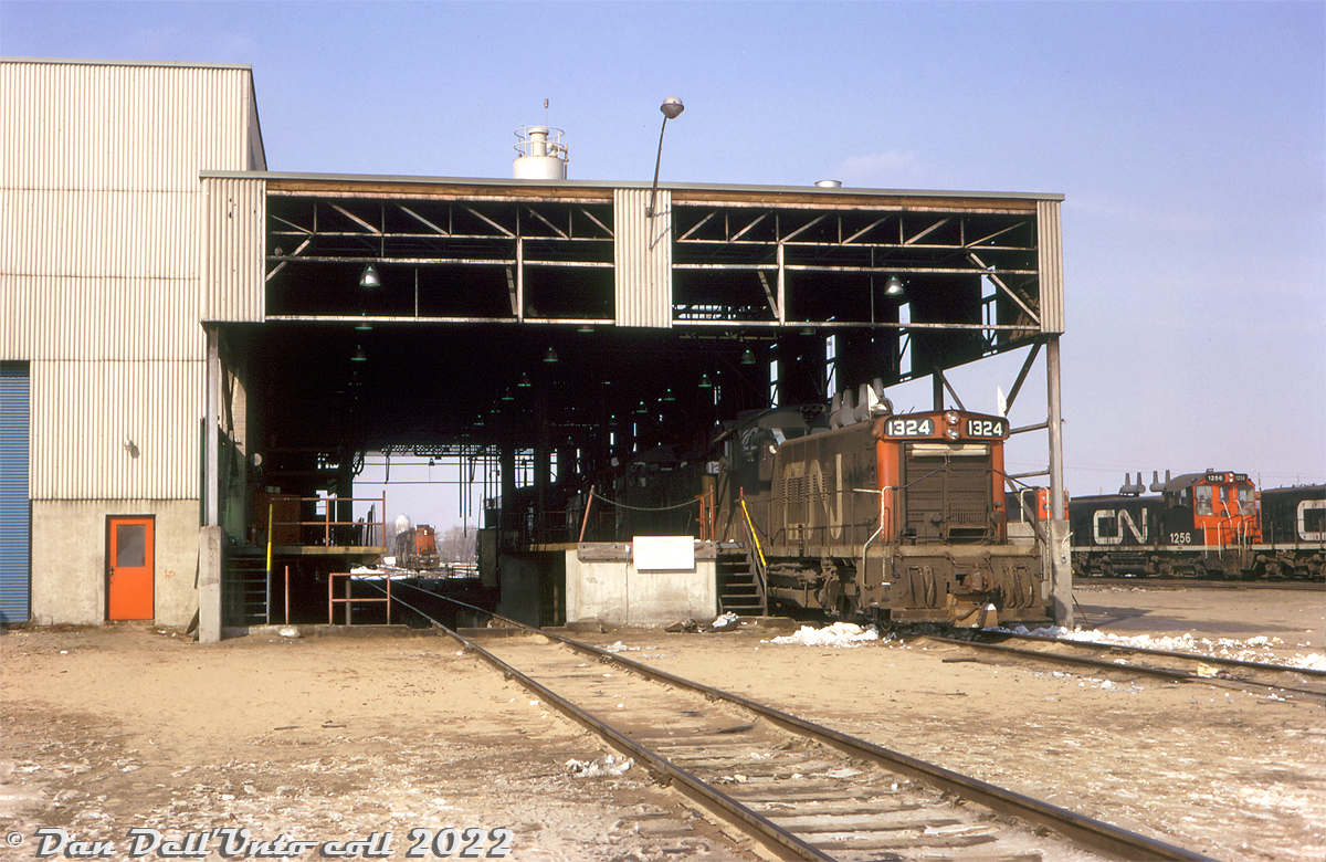 A group of CN SW1200RS units lead by 1324 (sporting white extra flags) sit under the canopy in the open servicing bays at the east end of Toronto Yard's diesel shop, awaiting attention from shop forces. Other units on the shop tracks include other SW1200RS, GP9, and GP38-2 models - typical roadswitcher and local power at the time.  A 1200-series unit still in the old green and gold paint is visible under the canopy behind 1324, one of many units that evaded the new "noodle" logo and paint into the early 70's.

CN's Toronto Yard (later MacMillan Yard) diesel shop was originally built near the north-west end of the yard in the mid-1960's, and was expanded over time. The canopy area here was extended and enclosed sometime in the 1980's.

Original photographer unknown, Dan Dell'Unto collection slide.