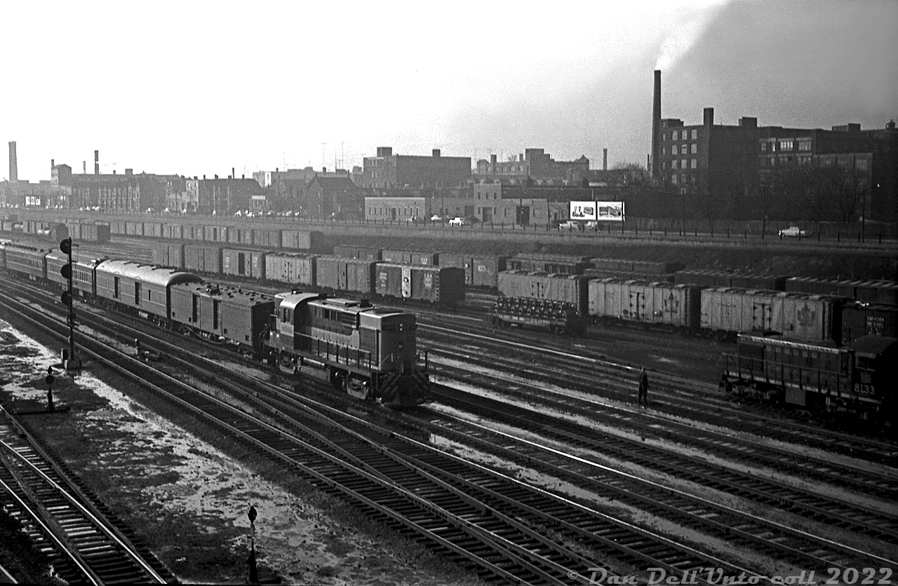 A steam-era view from the Spadina Avenue bridge looking west: CP RS10 8566 leads an inbound passenger train into downtown Toronto bound for Union Station, passing CN's Bathurst Street freight yards (full of the typical 50's freight car mix dominated by 40' boxcars and ice reefers, most sporting CN's maple leaf logo). An old 40' flatcar loaded with wheels is visible in the middle of the yard. Nearby, CN S2 8133 sits on one of the yard leads between switching duties.

The ever-present Front Street runs across the background, elevated above the yard, and populated by factories and houses that largely vanished in the following decades (a small gathering of historic houses along Draper Street remain today). The old Bathurst Tool building is visible on the far left. Judging by the sun angle and snow on the ground, this may be an evening winter shot.

Original photographer unknown, Dan Dell'Unto collection negative.