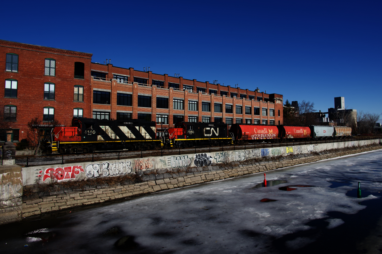 GP9s CN 4140 & CN 7060 are shoving four grain loads towards Ardent Mills, visible at far right. The train is paralleling the defrosting Lachine Canal.