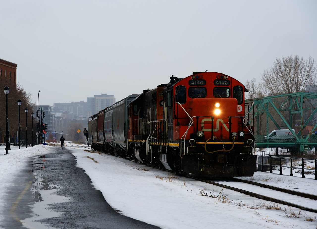 Light snow is falling as five grain loads are being shoved towards Ardent Mills by CN 4140 & CN 4720, with the conductor hanging onto the first car as they approach a busy crossing.