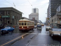 A rainy day in October 1973 finds TTC Peter Witt streetcar 2766 deadheading through mid-town Toronto, heading westbound on St. Clair Avenue crossing Yonge Street. The regular "Tour Tram" routing that 2766 was restored for months earlier ran around downtown streetcar trackage, so 2766 could be running a private fantrip coming from the nearby St. Clair subway station. A westbound PCC operating in regular service on the St. Clair route follows a few cars behind, and another PCC can be seen turning into St. Clair station's loop, probably a westbound from Eglinton Loop <a href=http://www.railpictures.ca/?attachment_id=32380><b>via Mount Pleasant</b></a>/St. Clair.
<br><br>
Large office towers have since replaced both the TD Bank building at the northeast and the Super Save Drug Mart at the southeast corners. License plate afficionados, note the brand new original A-series license plate on the right: 1973 was the first year Ontario plates weren't replaced on a yearly bases (rather, they required annual sticker updates to be valid).
<br><br>
<i>Original photographer unknown, Dan Dell'Unto collection slide.</i>