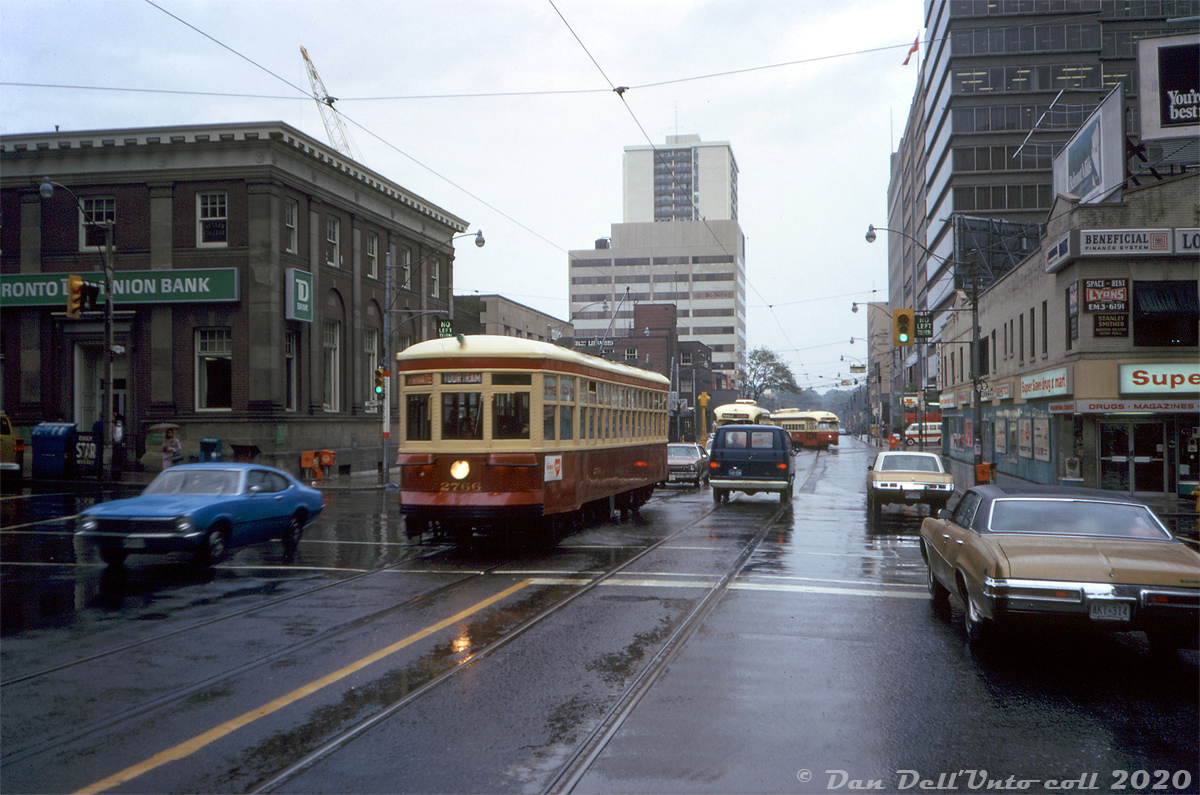 A rainy day in October 1973 finds TTC Peter Witt streetcar 2766 deadheading through mid-town Toronto, heading westbound on St. Clair Avenue crossing Yonge Street. The regular "Tour Tram" routing that 2766 was restored for months earlier ran around downtown streetcar trackage, so 2766 could be running a private fantrip coming from the nearby St. Clair subway station. A westbound PCC operating in regular service on the St. Clair route follows a few cars behind, and another PCC can be seen turning into St. Clair station's loop, probably a westbound from Eglinton Loop via Mount Pleasant/St. Clair.

Large office towers have since replaced both the TD Bank building at the northeast and the Super Save Drug Mart at the southeast corners. License plate afficionados, note the brand new original A-series license plate on the right: 1973 was the first year Ontario plates weren't replaced on a yearly bases (rather, they required annual sticker updates to be valid).

Original photographer unknown, Dan Dell'Unto collection slide.