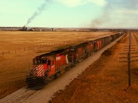 Westward from Medicine Hat, CP climbs out of the South Saskatchewan River valley to Redcliff and crosses under highway 1 at mile 3.8, my photo perch on Monday 1974-09-30 for train No. 965 with “four nickels” SD40 5555 and C-424 4248 and C-636 4728, with the petrochemical facility at Cousins in the distance.  Today, no poles and no fence.